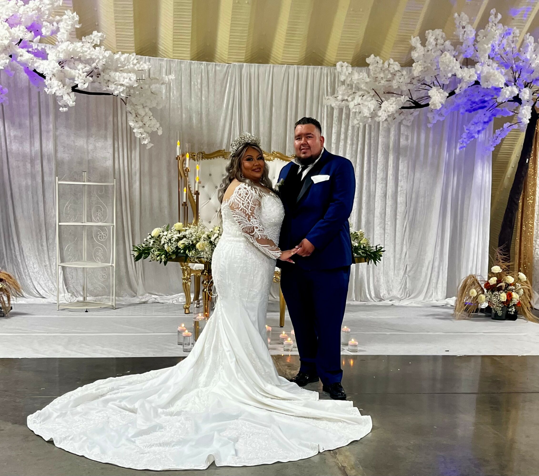 Bride and groom holding hands on stage with white floral decorations and draped backdrop behind them.