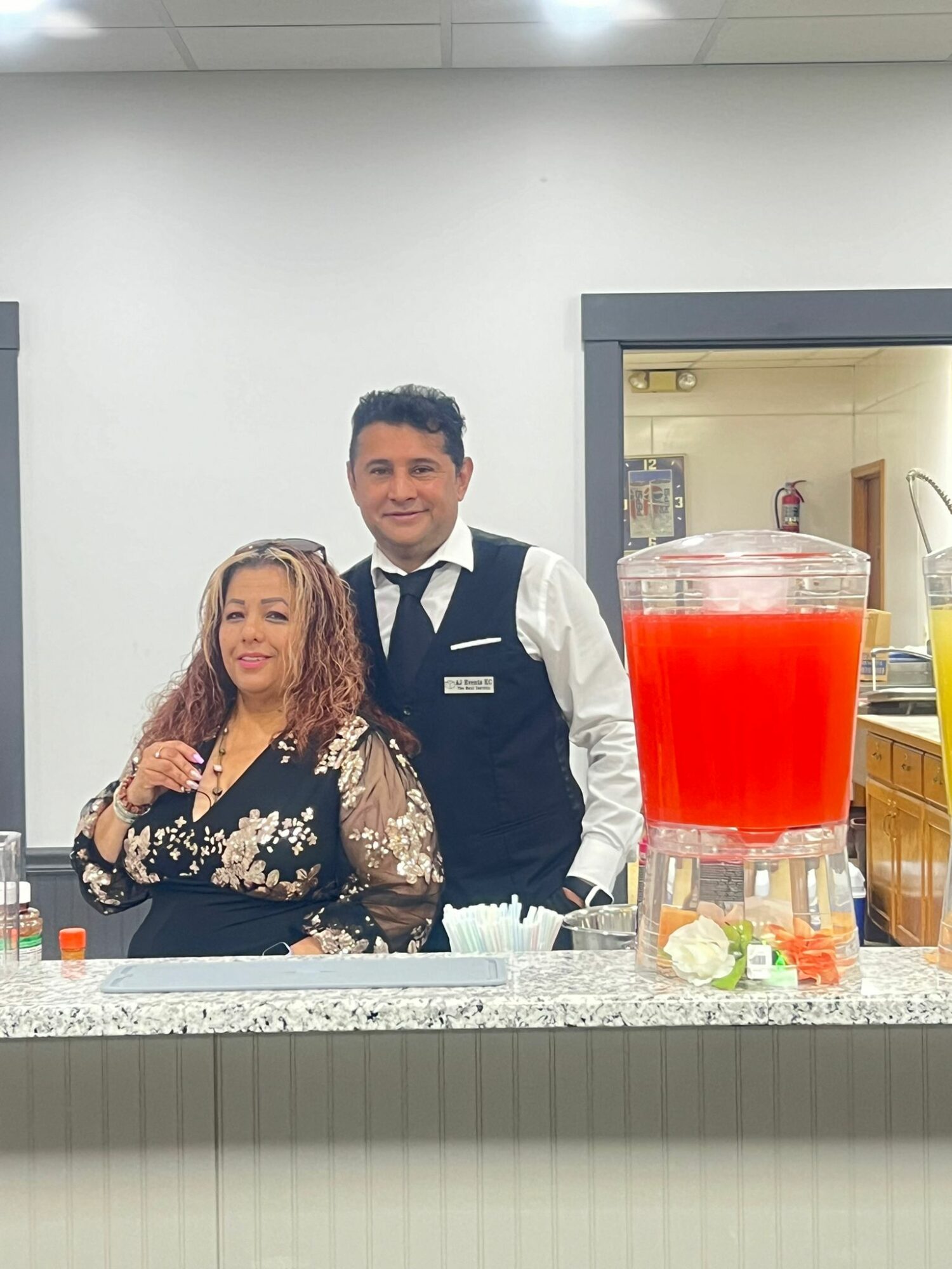 Two people standing behind a counter with a large red beverage dispenser, smiling, in an indoor setting.