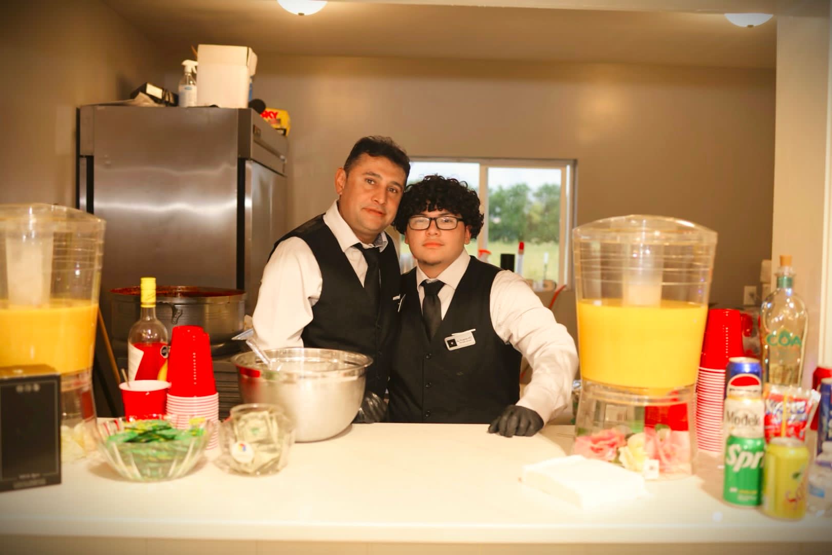 Two people behind a bar counter with drinks and supplies, one with glasses and curly hair, the other with short hair, in a room with a window.