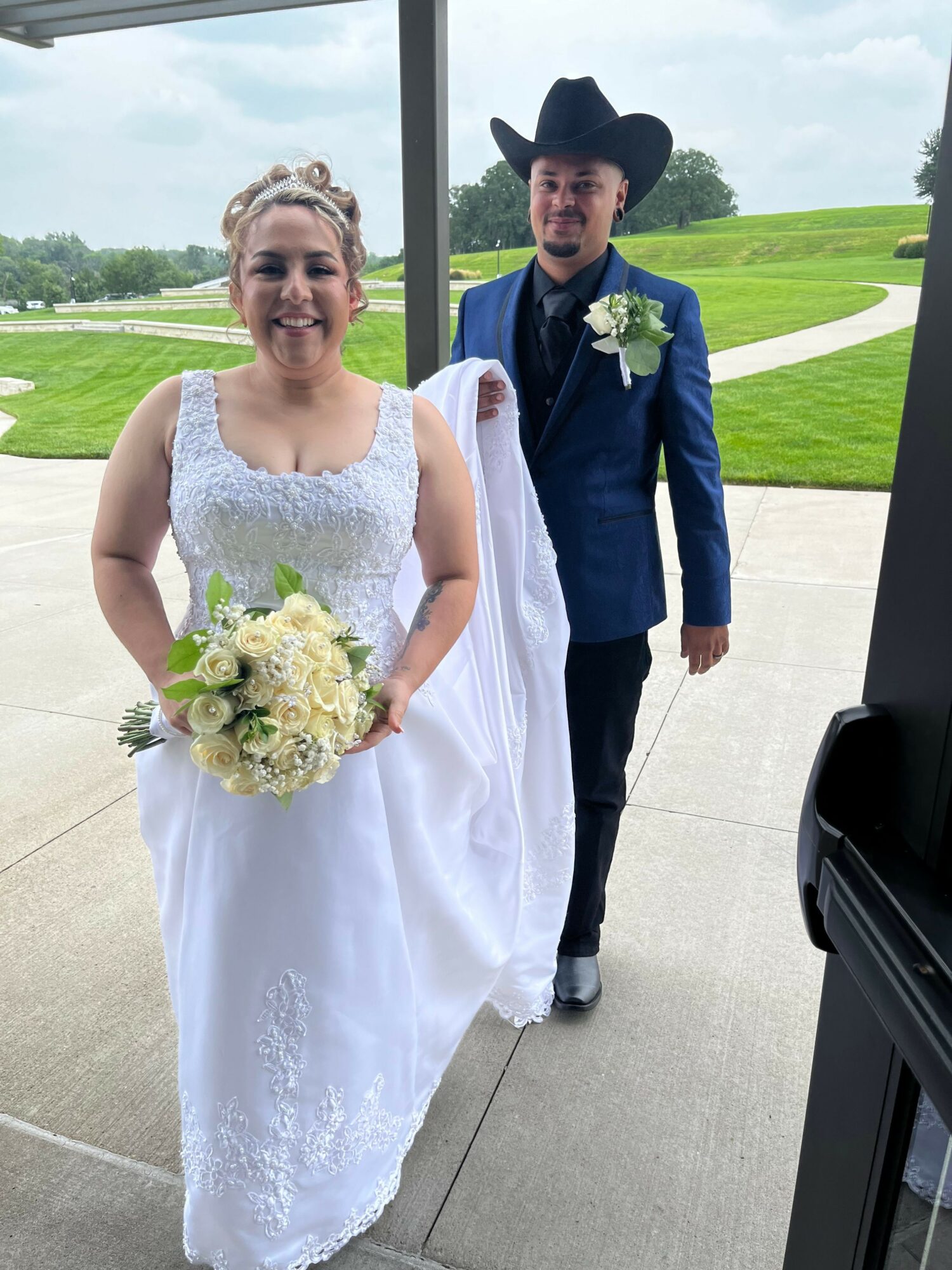Bride in white wedding dress holding bouquet, groom in blue suit and hat standing outside near a car, green landscape background.