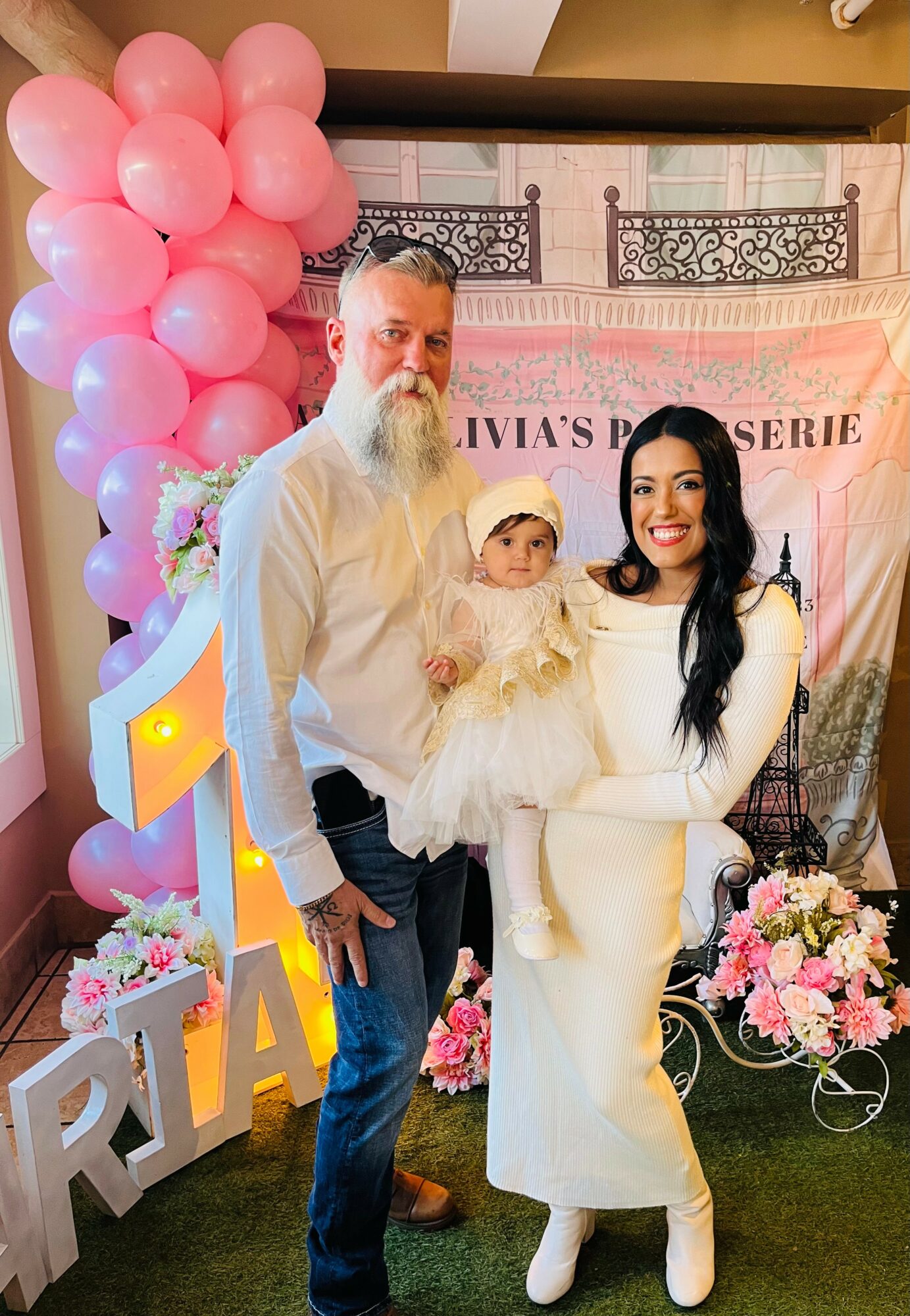 Three people, a man, woman, and child, stand in front of pink balloons and floral decorations at an event, smiling.