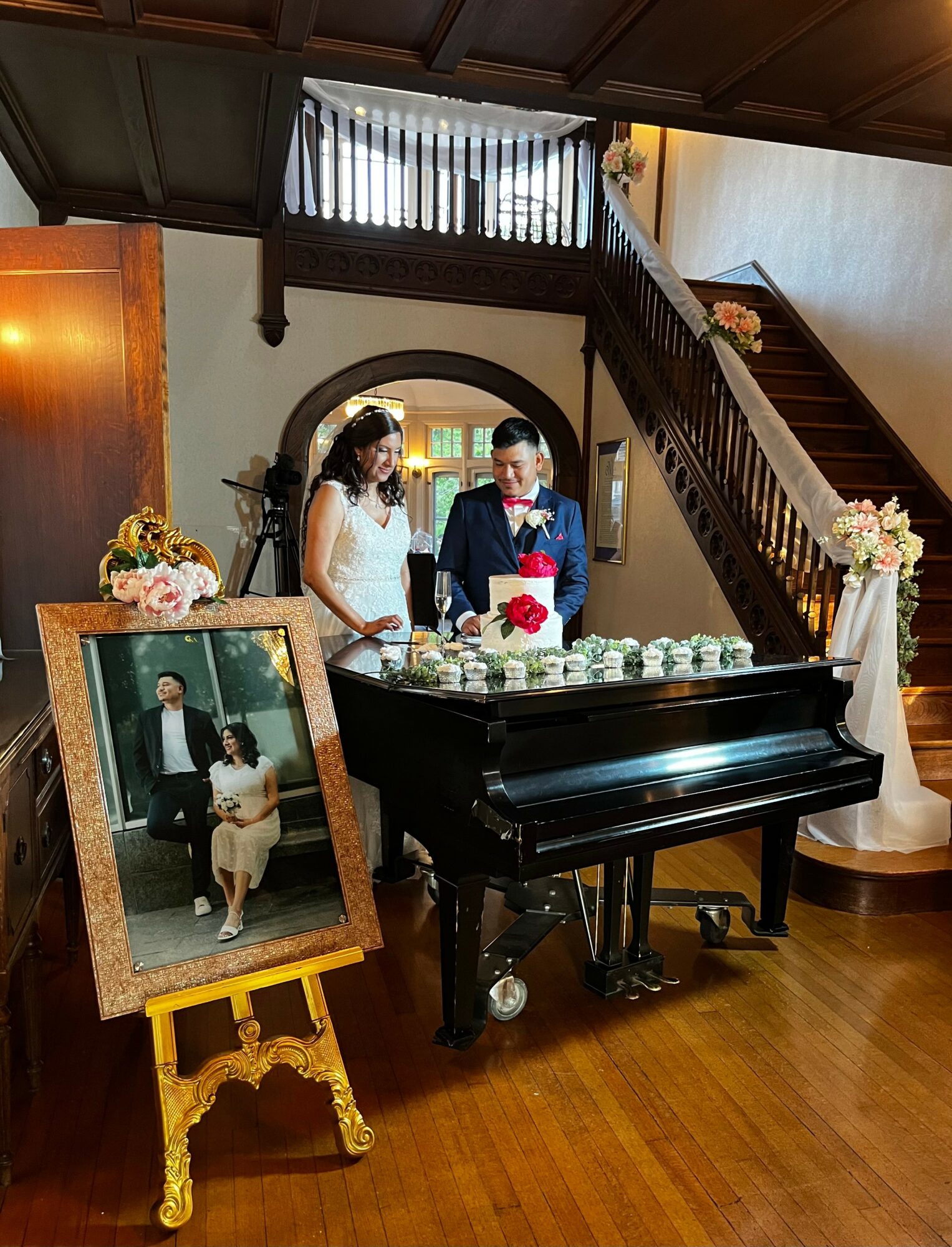 Couple in wedding attire standing behind a black piano with a framed photo on a yellow stand nearby, inside a wooden interior.