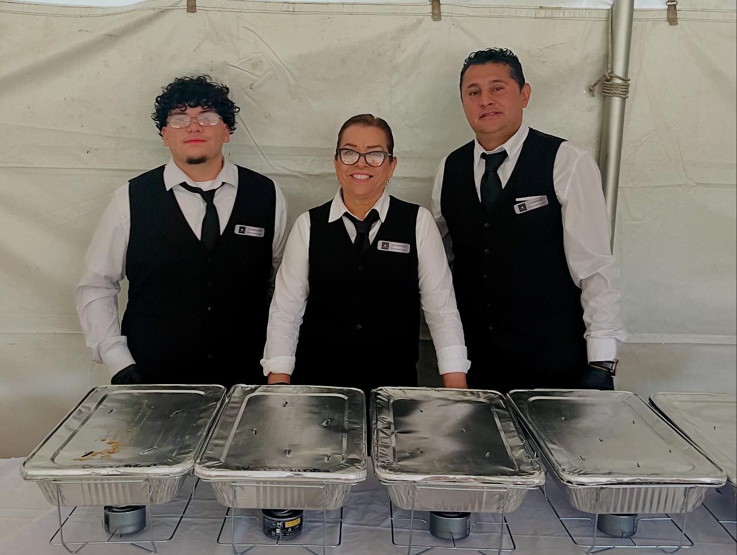 Three people in formal attire standing behind four covered serving trays inside a tent.