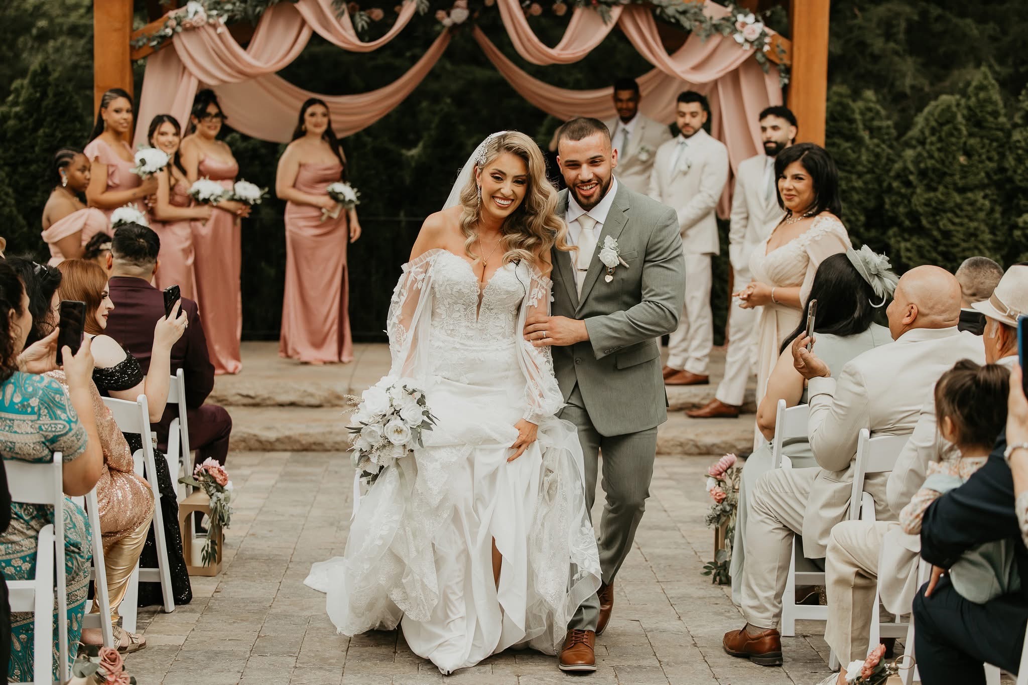 Bride and groom walking down aisle at outdoor wedding ceremony with guests seated on sides, floral decorations, and a draped backdrop.