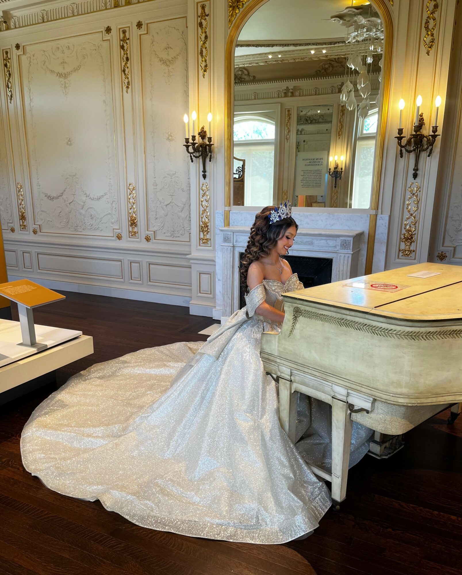 Young girl in a white dress with a long train, sitting at a grand piano in an ornate room with gold accents and mirrors.