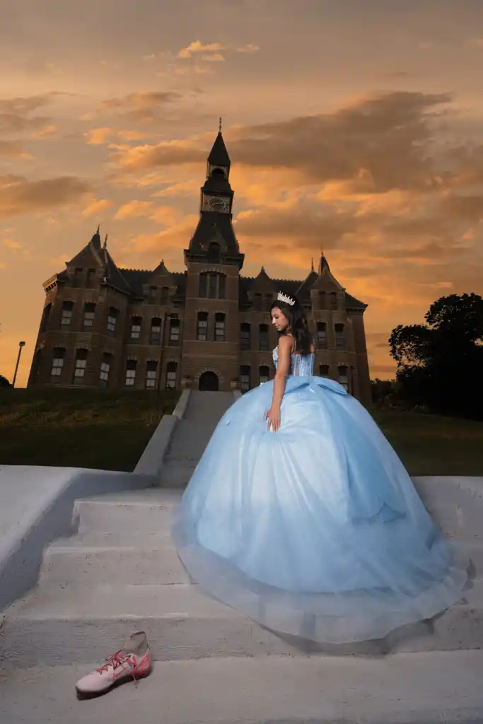 Young woman in a blue ball gown standing on stairs in front of a castle at sunset.