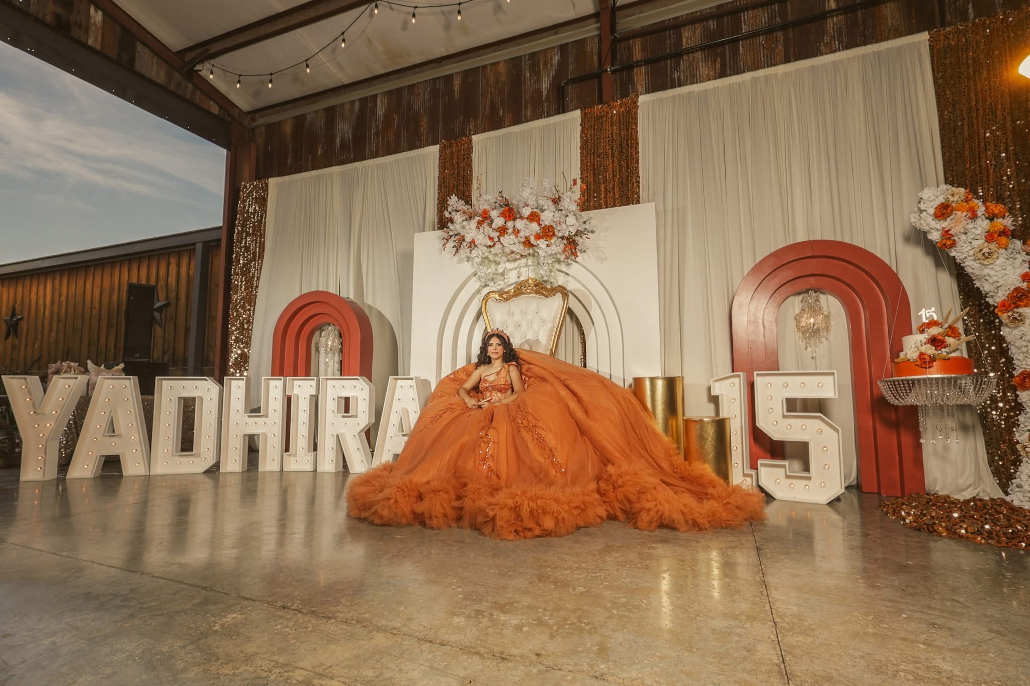 Person in an orange gown sitting on the floor in front of large decorative numbers and letters, with floral arrangements and curtains behind.