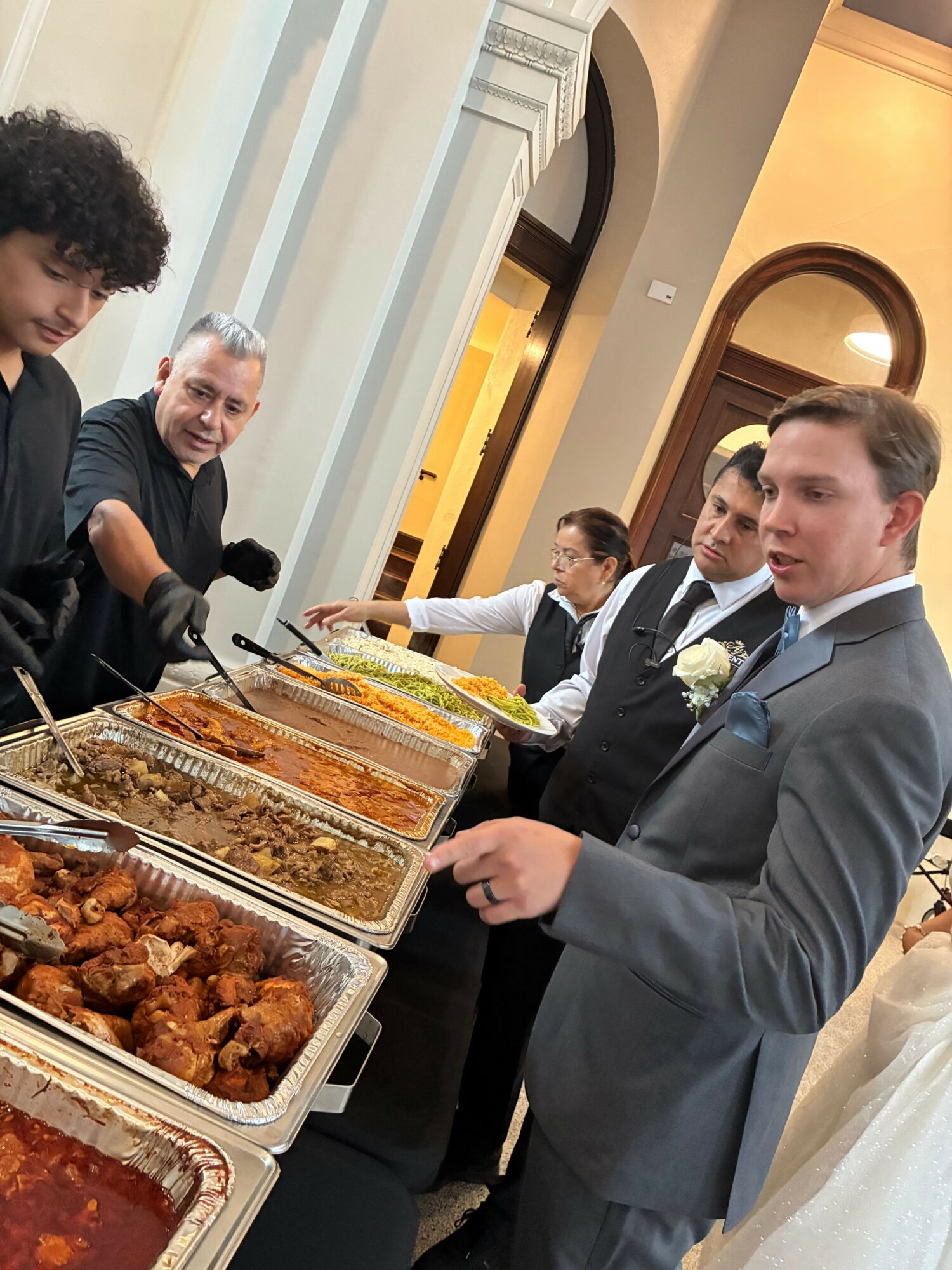 People serving food at a buffet table during a formal event, with a man in a suit and others in formal attire.