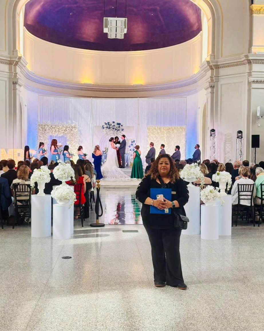 Woman standing in front of a wedding ceremony with guests and a decorated stage inside a grand hall.