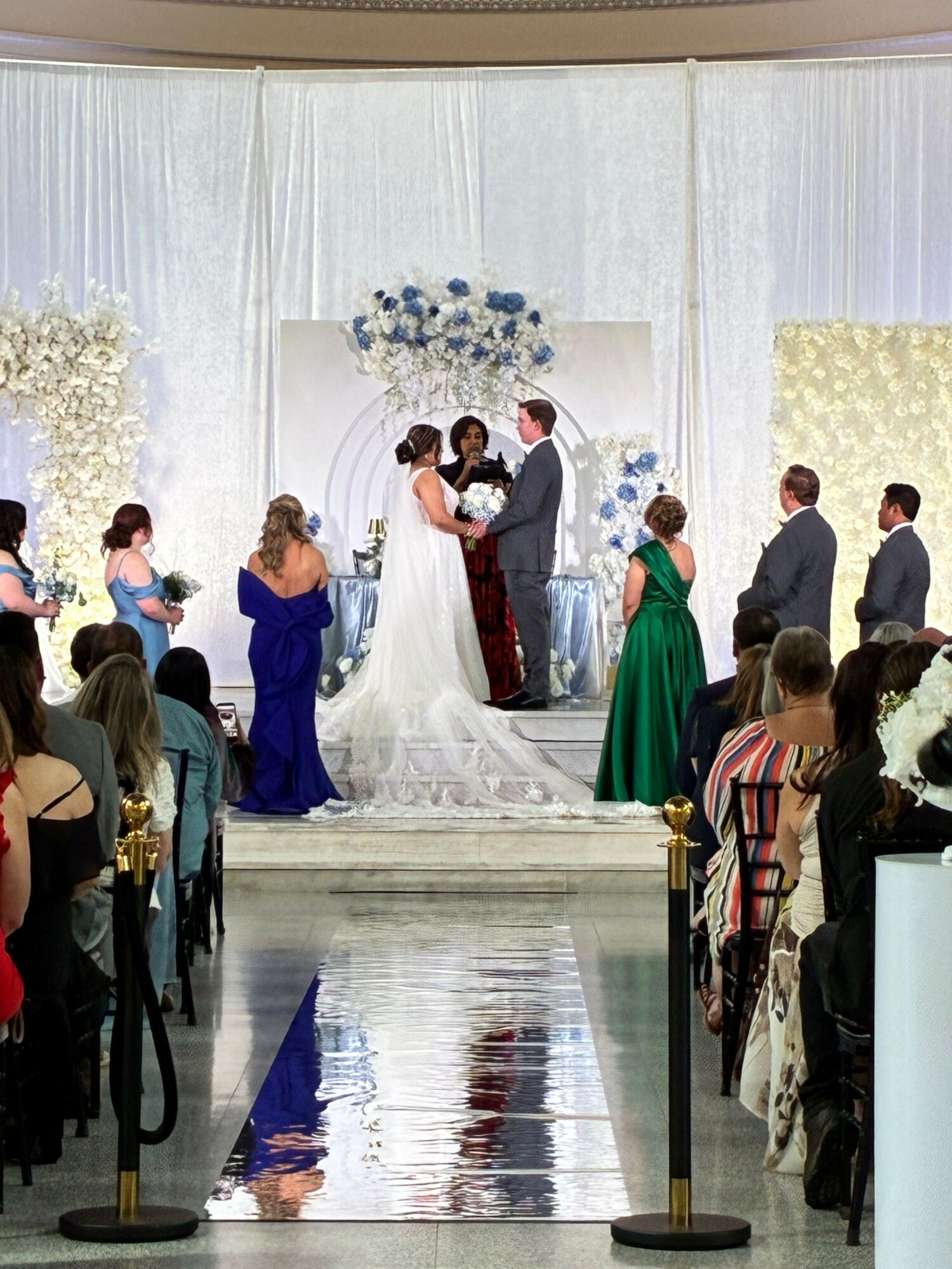 People gather around a wedding cake at a celebration, with floral decorations and a white backdrop.