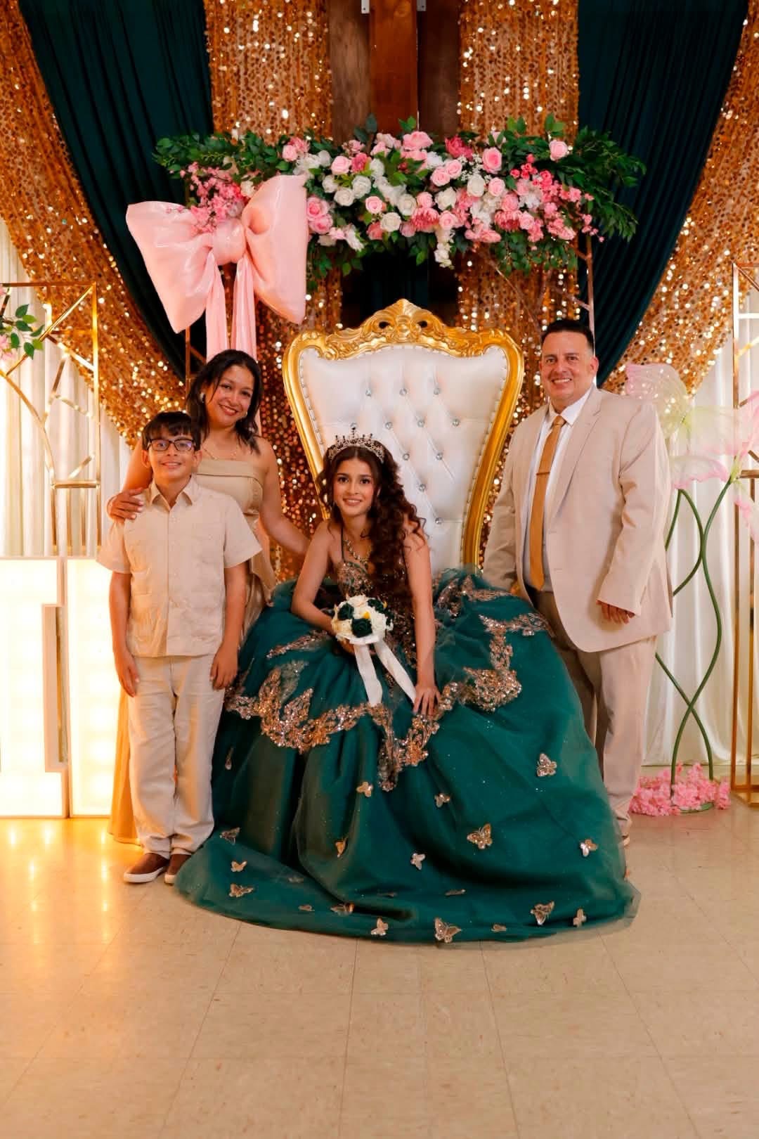 A girl in a green dress sits on a white ornate throne with a man and two women standing beside her, decorated with pink flowers and gold curtains.