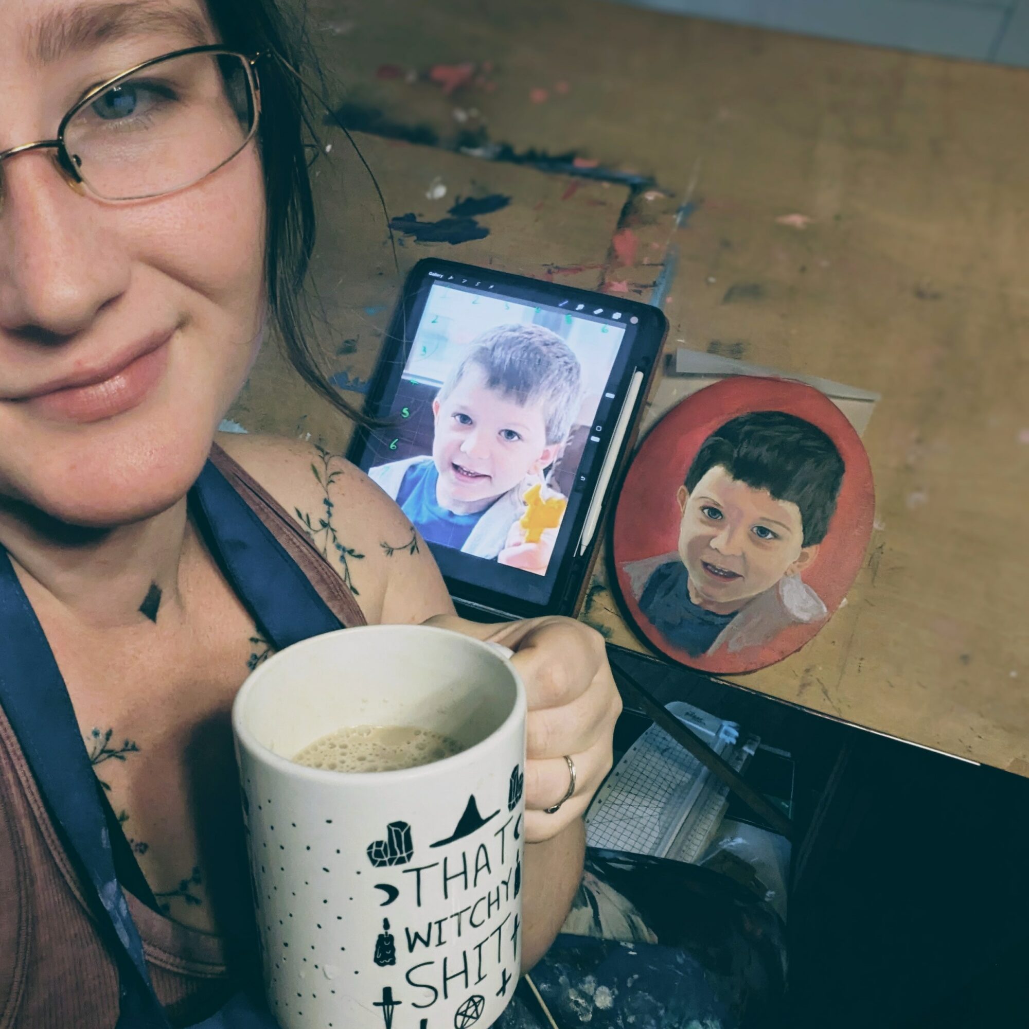Person holding a mug with text, with a tablet showing a child's photo and a painted portrait of the same child on a table.