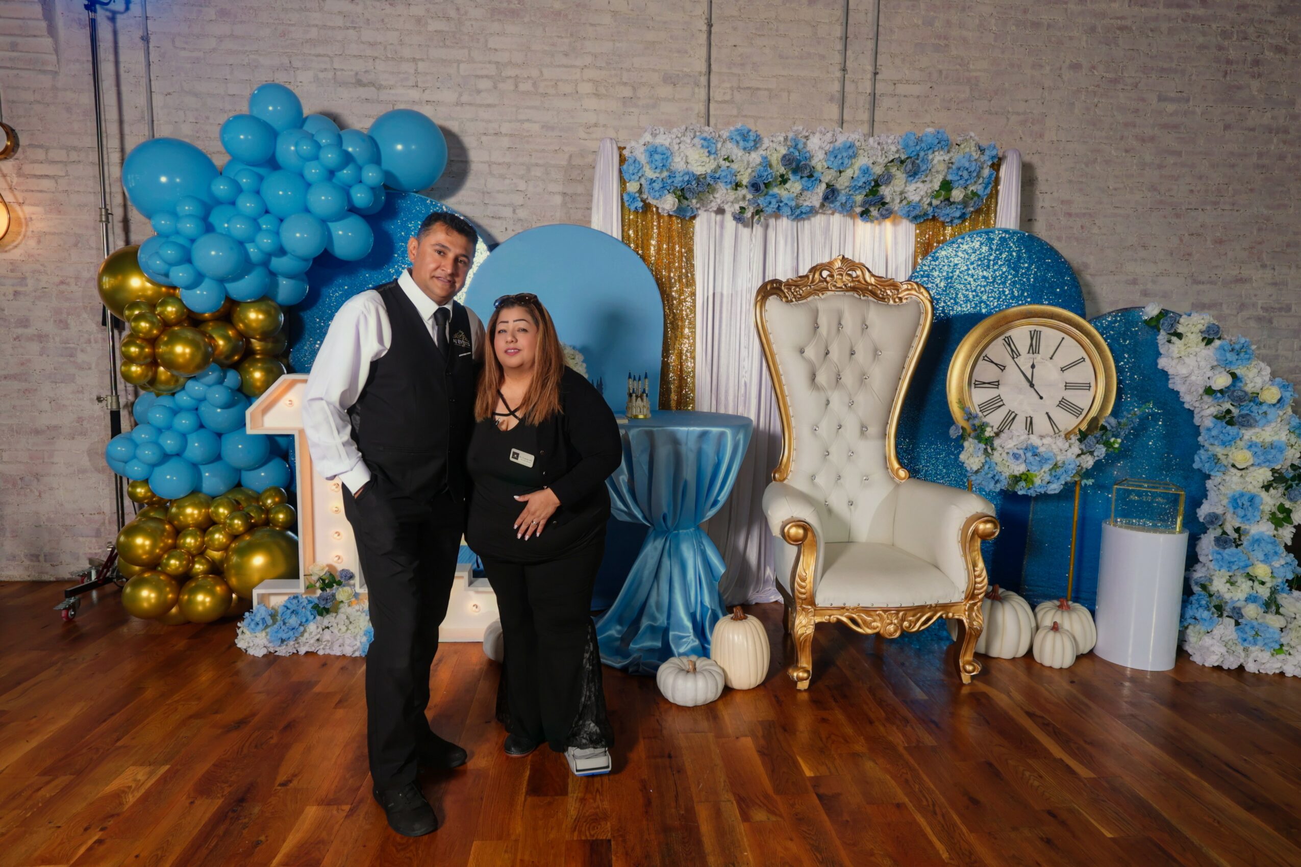 Two people stand in front of a decorated backdrop with a large white and gold chair, blue and white flowers, and balloons.