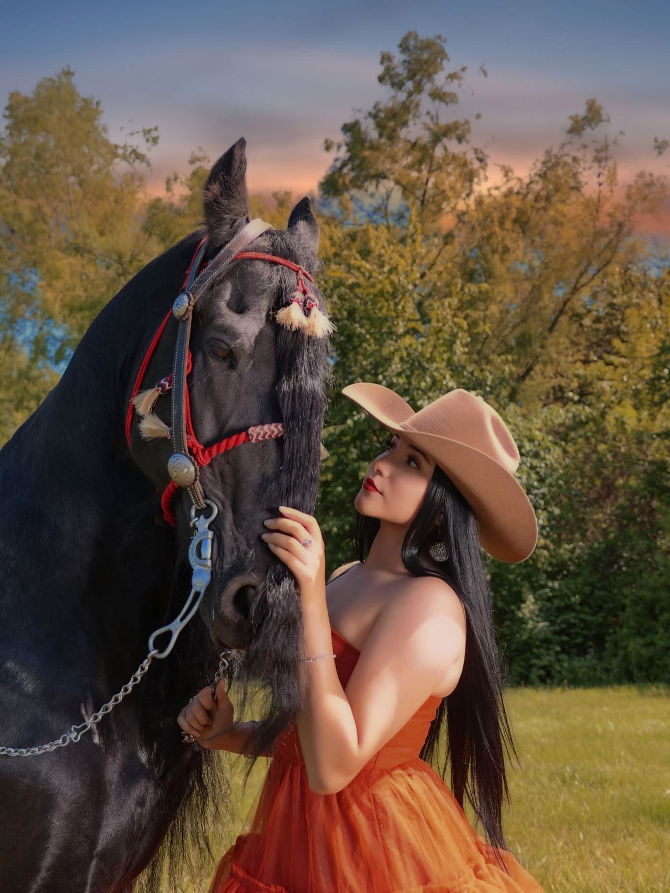 Young woman in a wide-brimmed hat gently touches a black horse outdoors with trees and a sunset sky in the background.