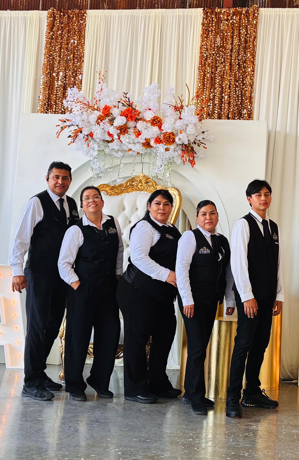 Five people in formal attire standing in front of a decorated backdrop with white and gold curtains and a floral arrangement.
