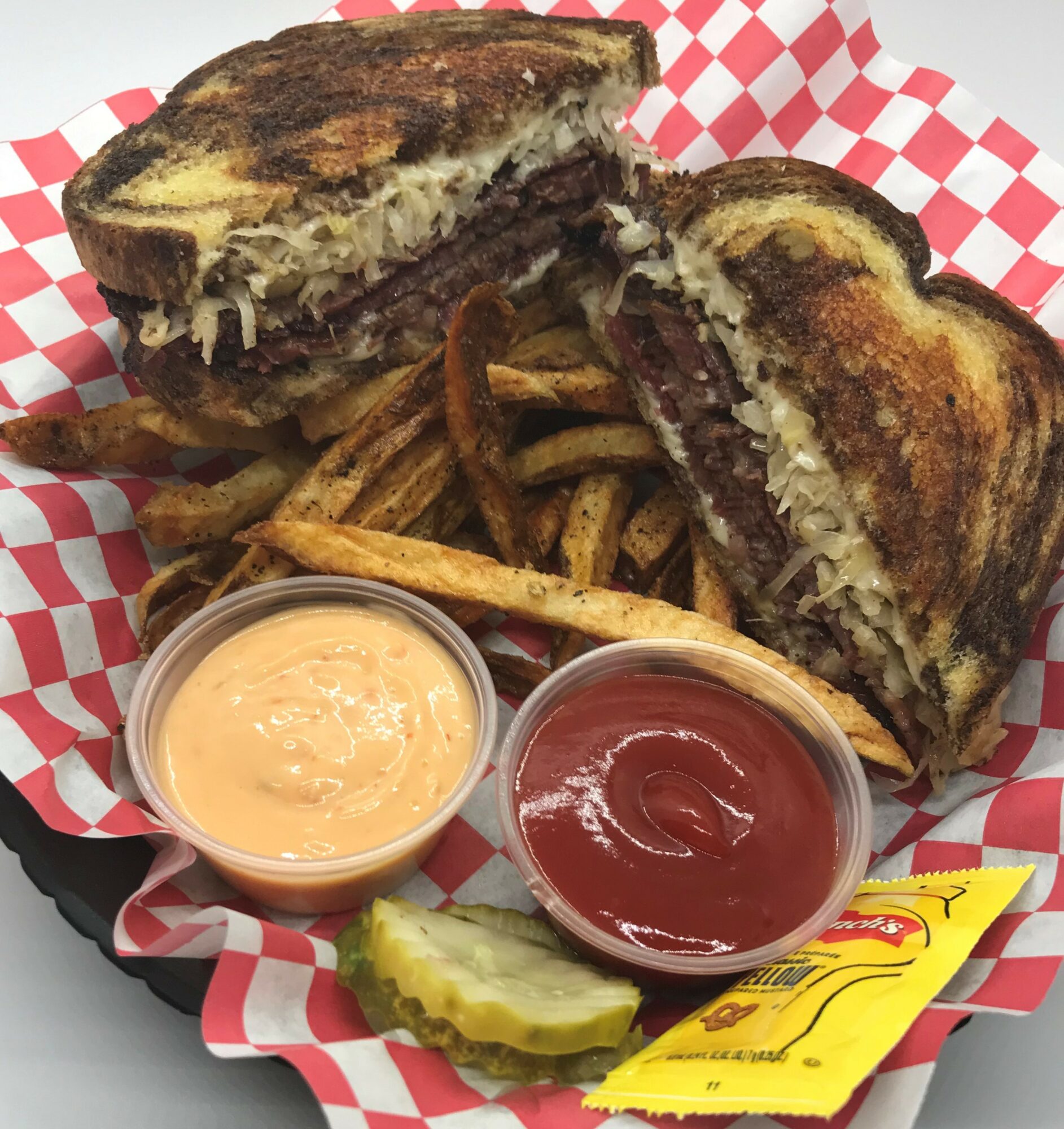 Grilled sandwich with fries, pickles, and condiments on red and white checkered paper.