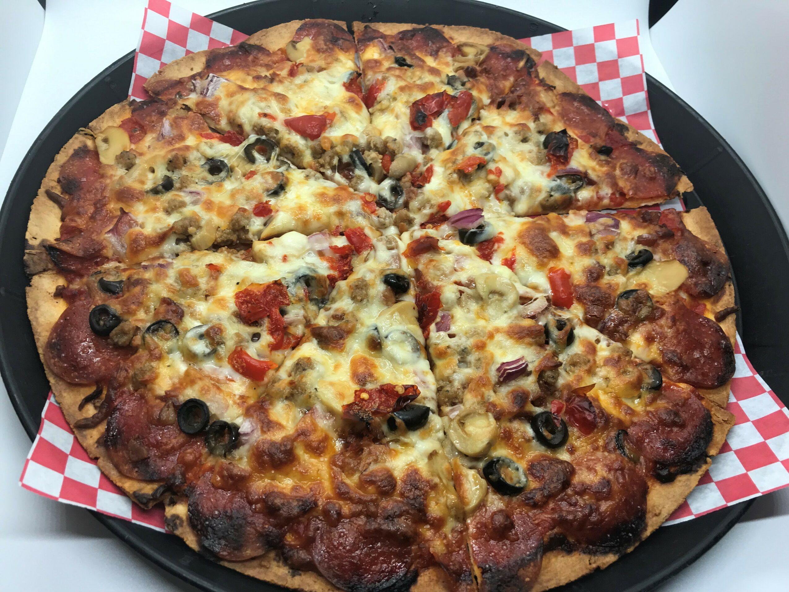 Pizza with cheese, black olives, red peppers, and meat on a black tray with red and white checkered paper, viewed from above.