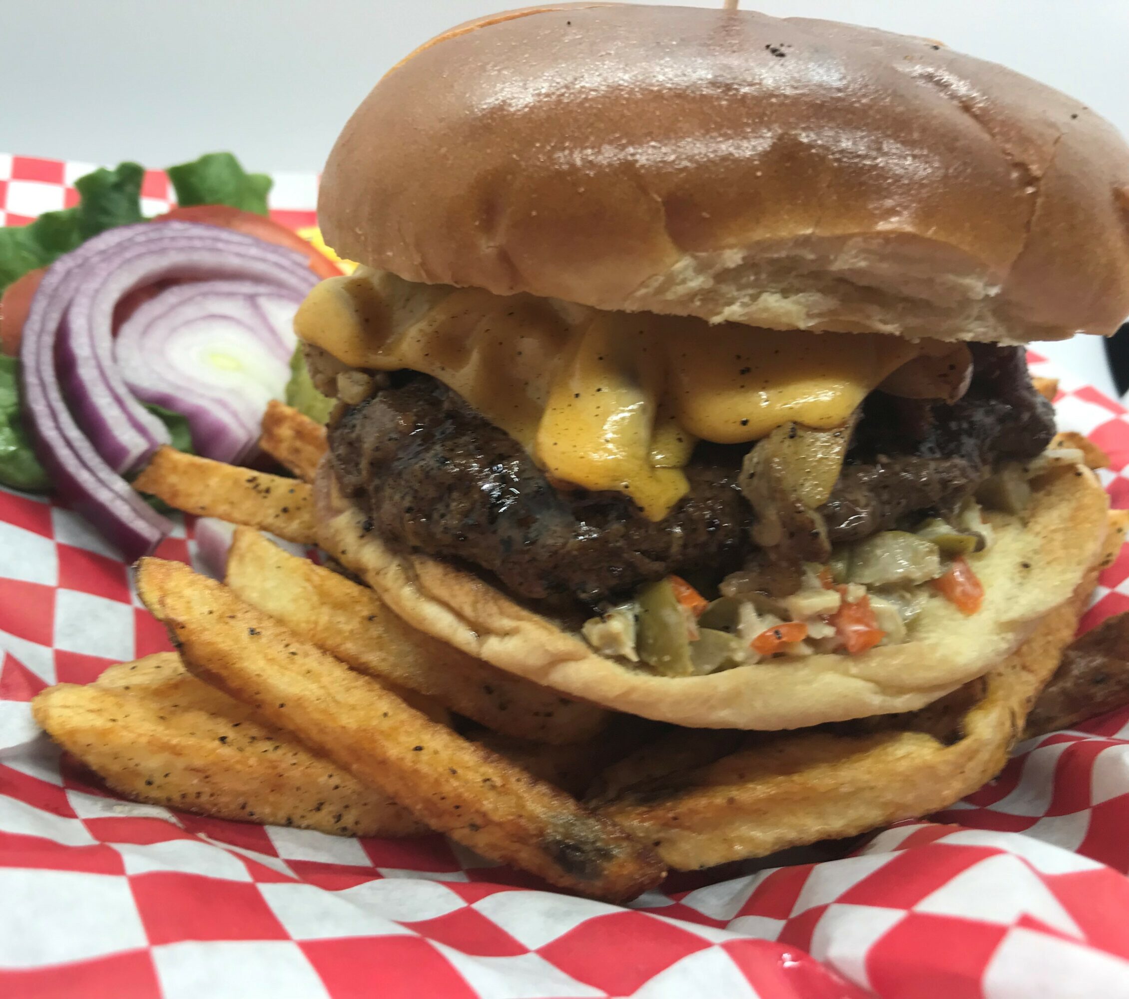 Burger with cheese, beef patty, pickles, onions, and sauce, served with fries on red and white checkered paper.