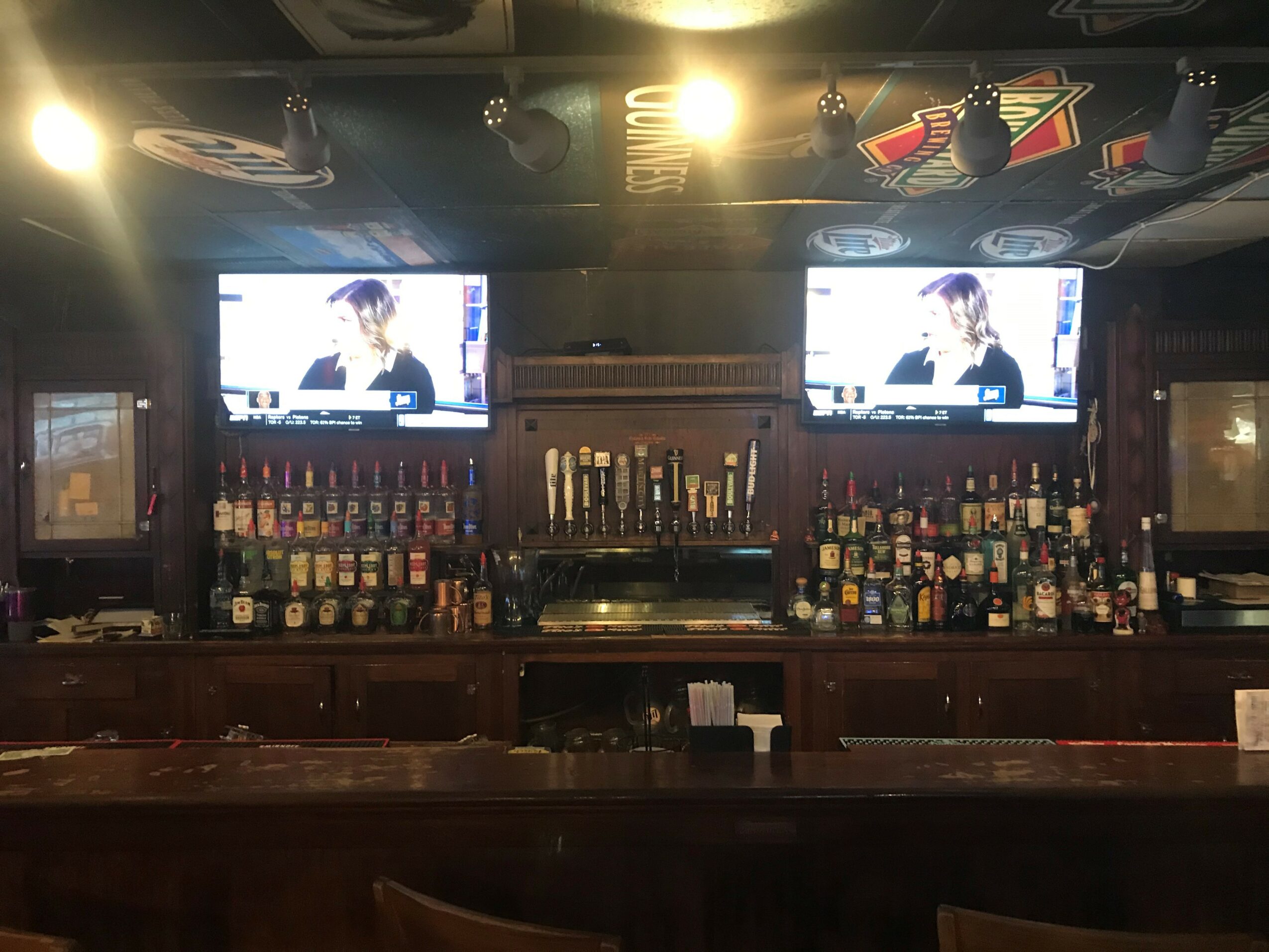 Bar with two TV screens, shelves with bottles, and a dark wooden counter.
