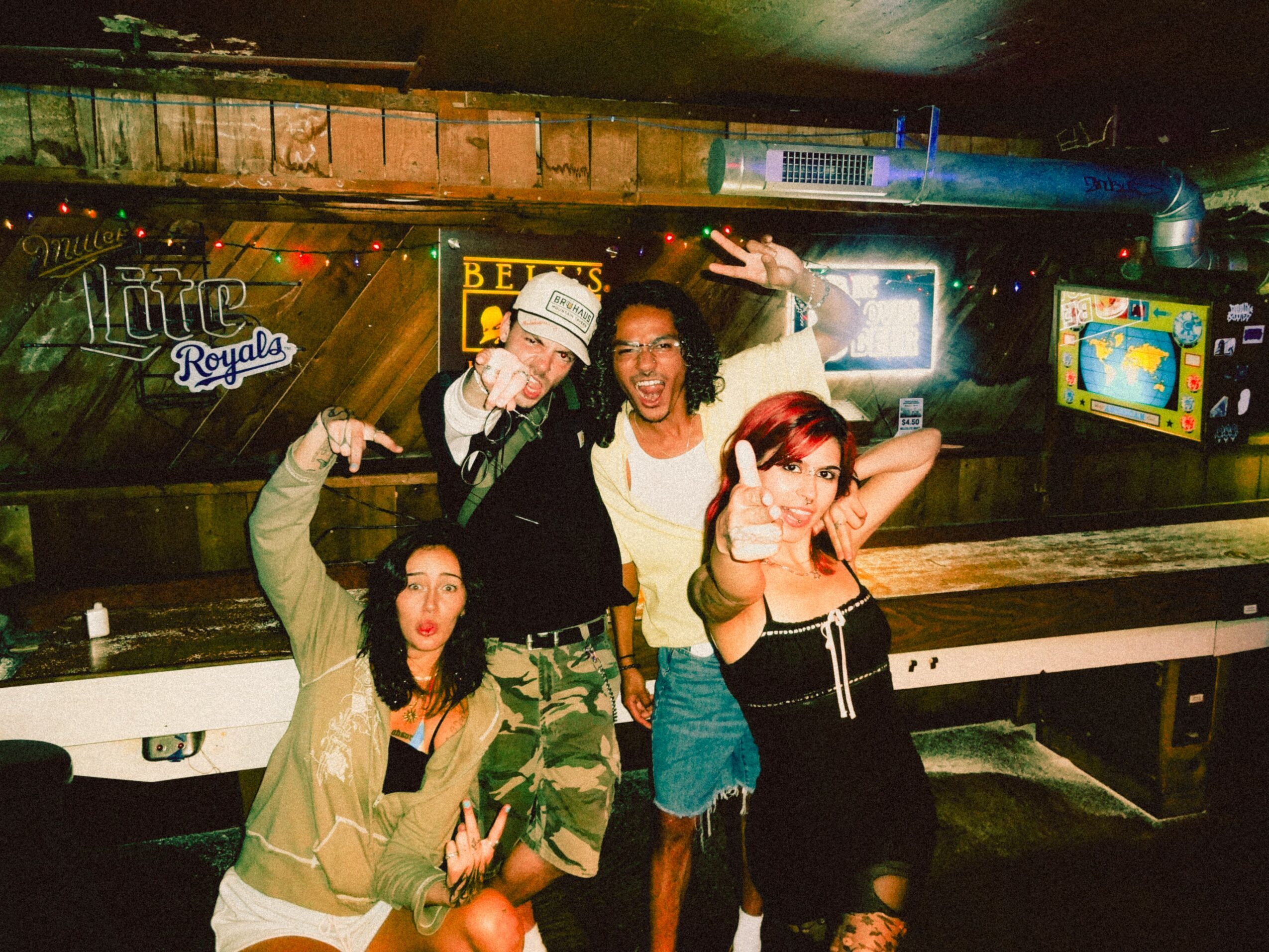 Group of four people posing in a bar with neon signs and TV screens in the background.