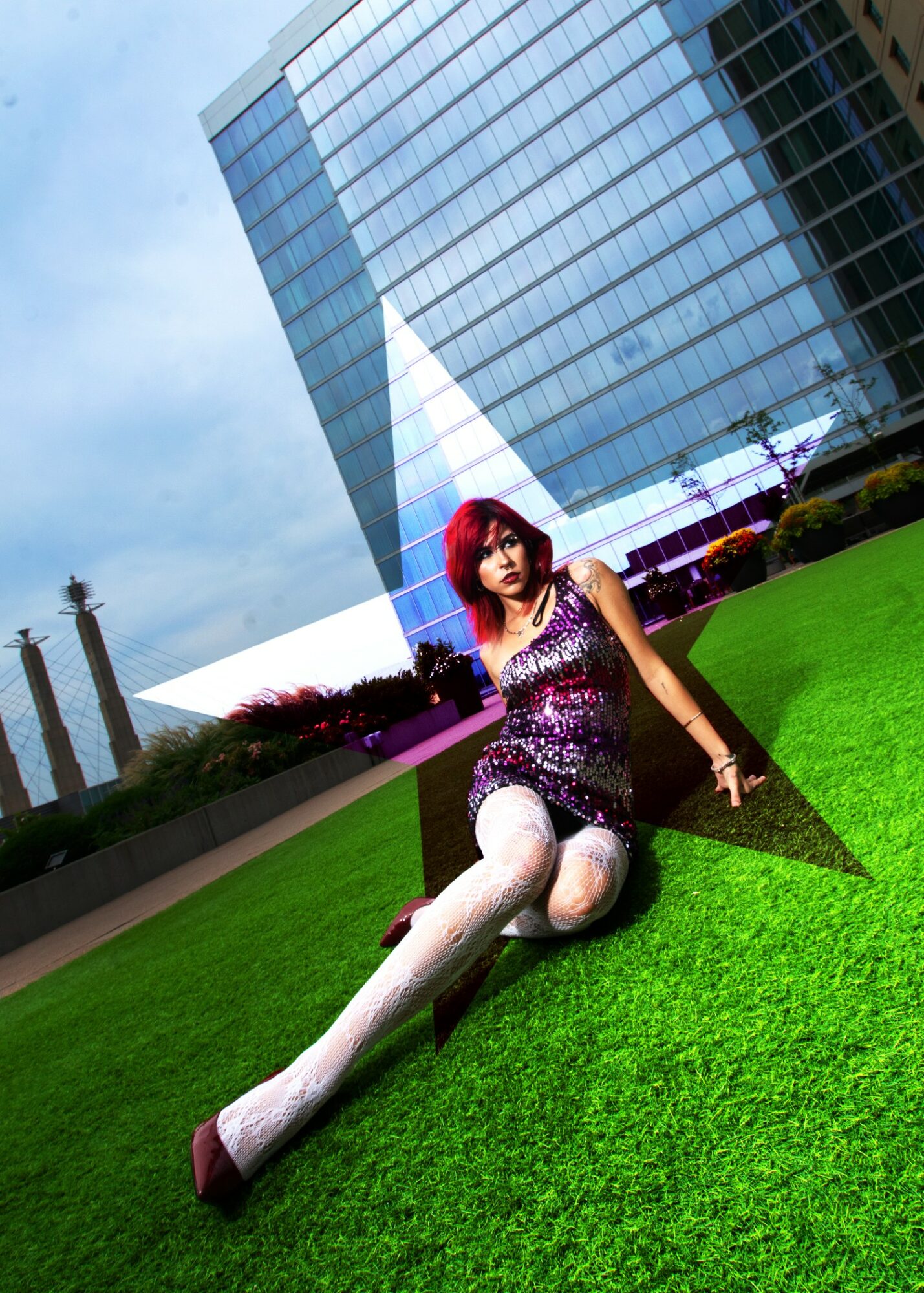 Woman with red hair sitting on green grass in front of a modern glass building, under a cloudy sky.