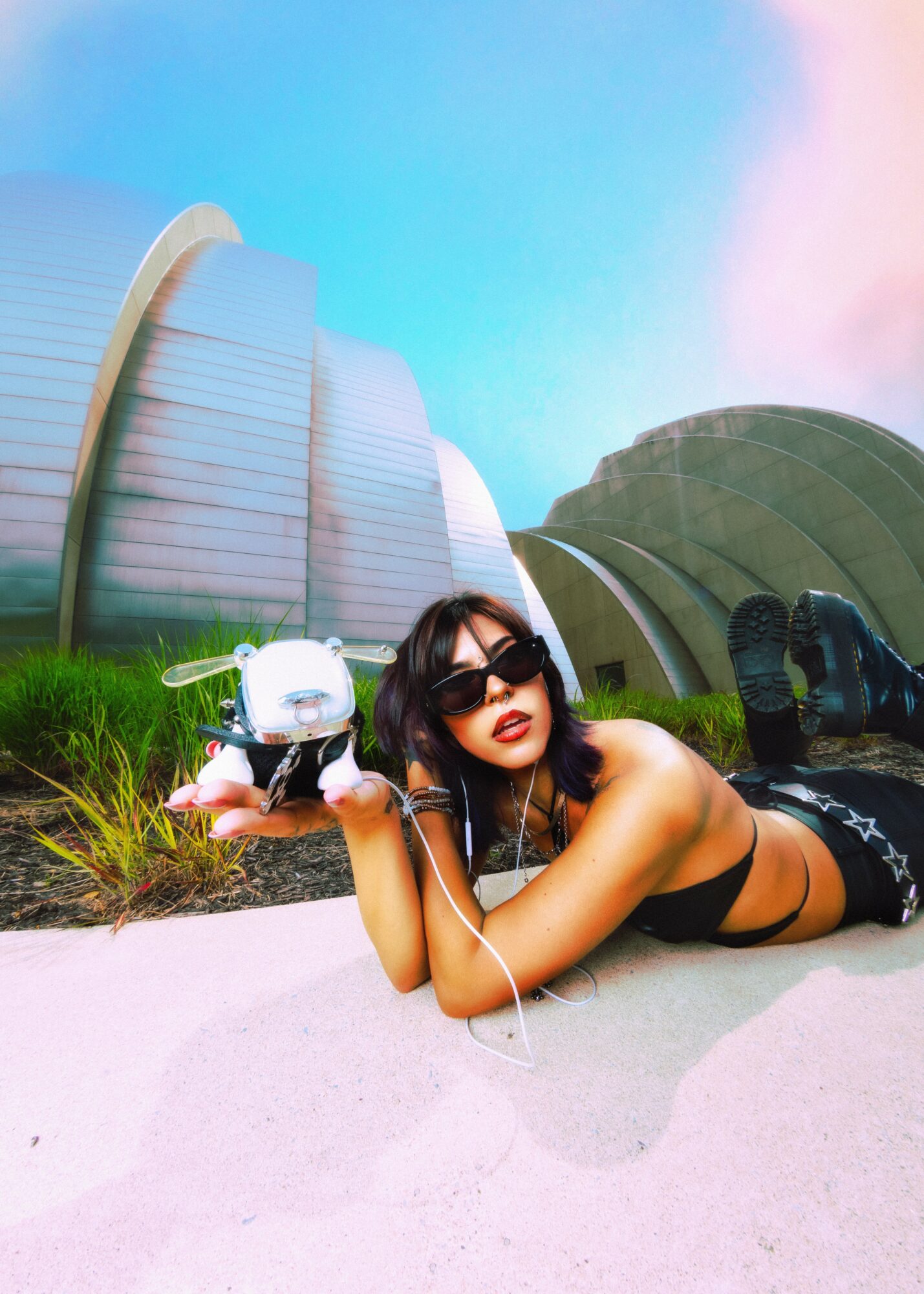 Woman lying on sand holding a camera, wearing sunglasses and black clothing, with modern buildings in the background.
