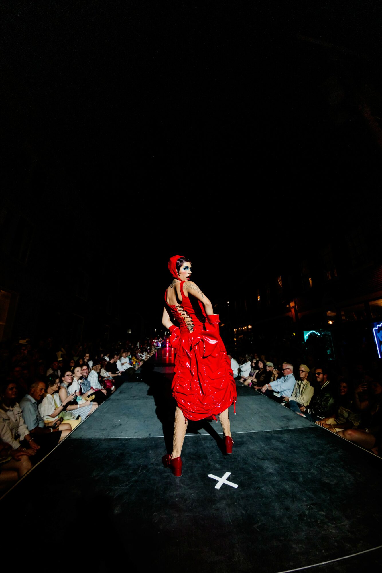 Performer in red dress and high heels on stage with audience in background, dark lighting, spotlight on performer.