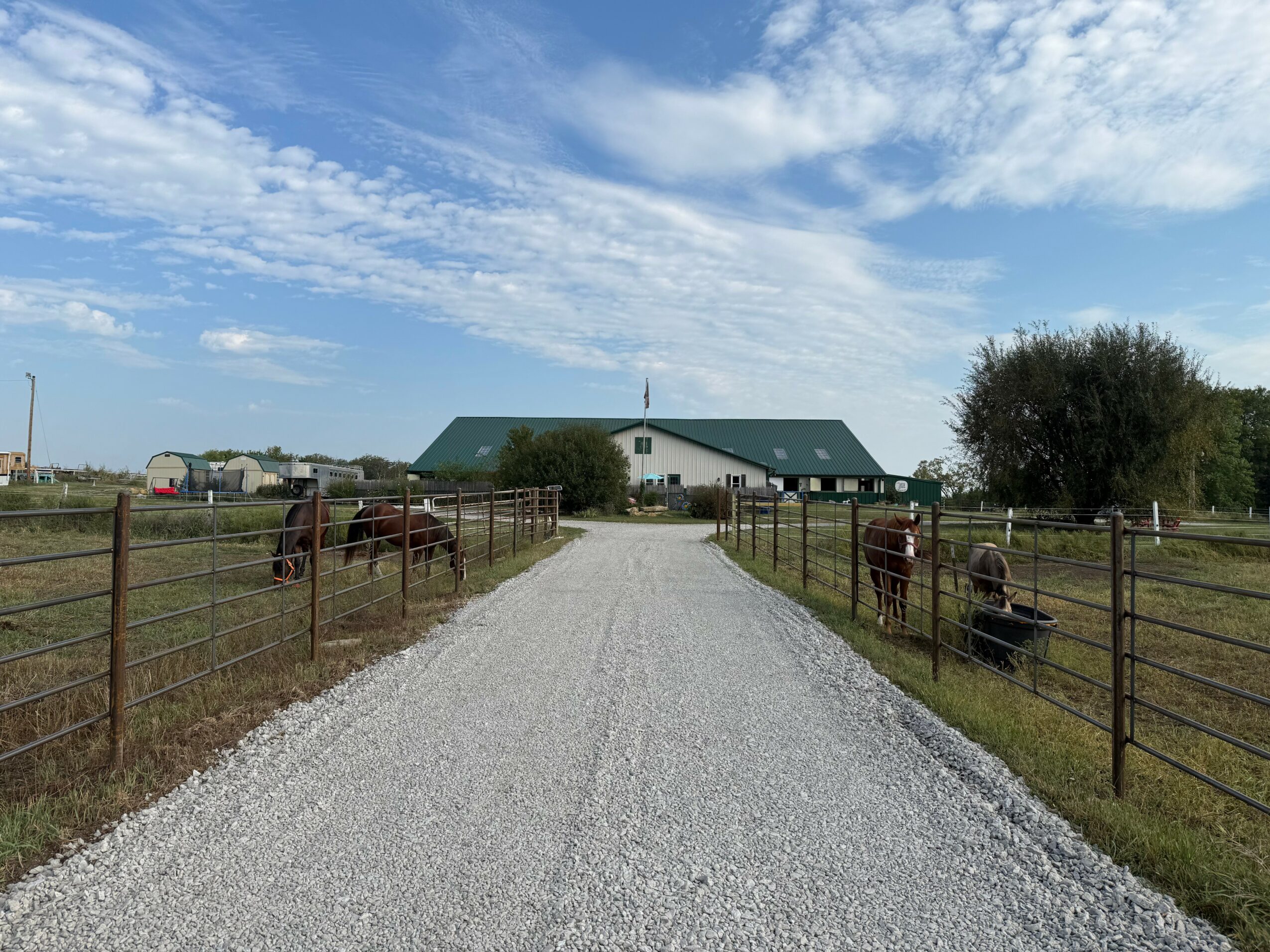 Gravel driveway leading to a white building with a green roof, surrounded by fenced grassy fields and trees, under a partly cloudy sky.