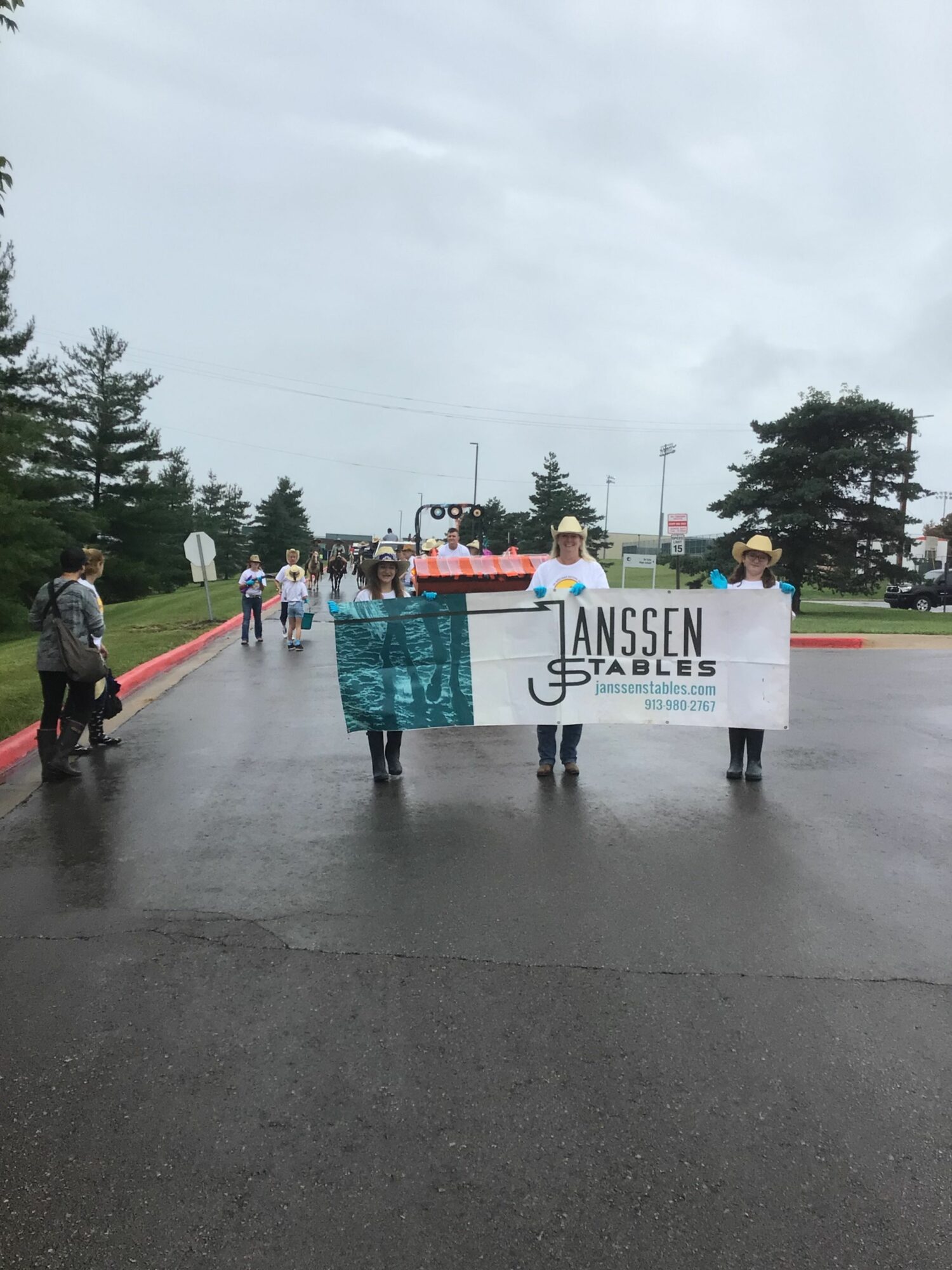 People gather around a table with a banner at an outdoor event, overcast sky, trees in background.