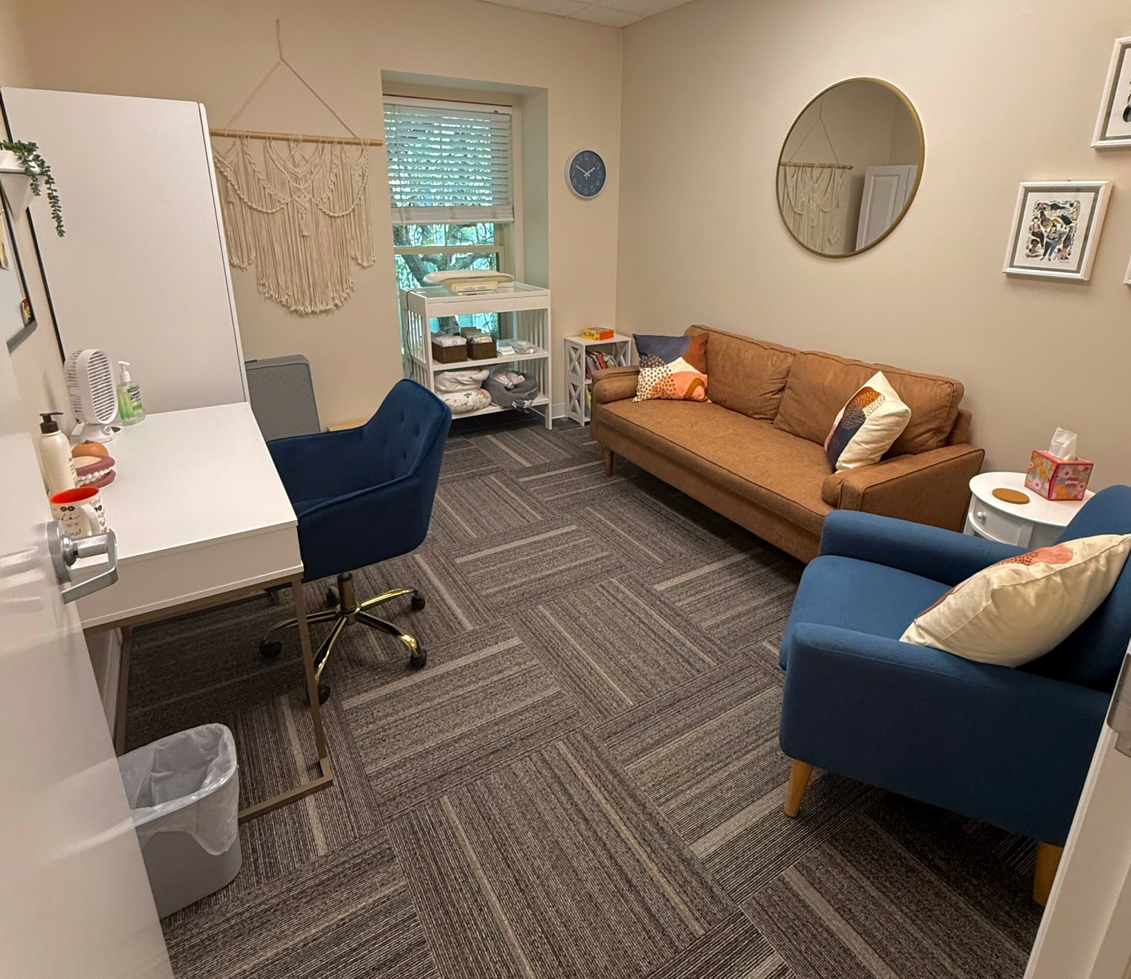 Living room with a brown sofa, blue armchair, white desk, and window with blinds, decorated with wall art and a mirror.
