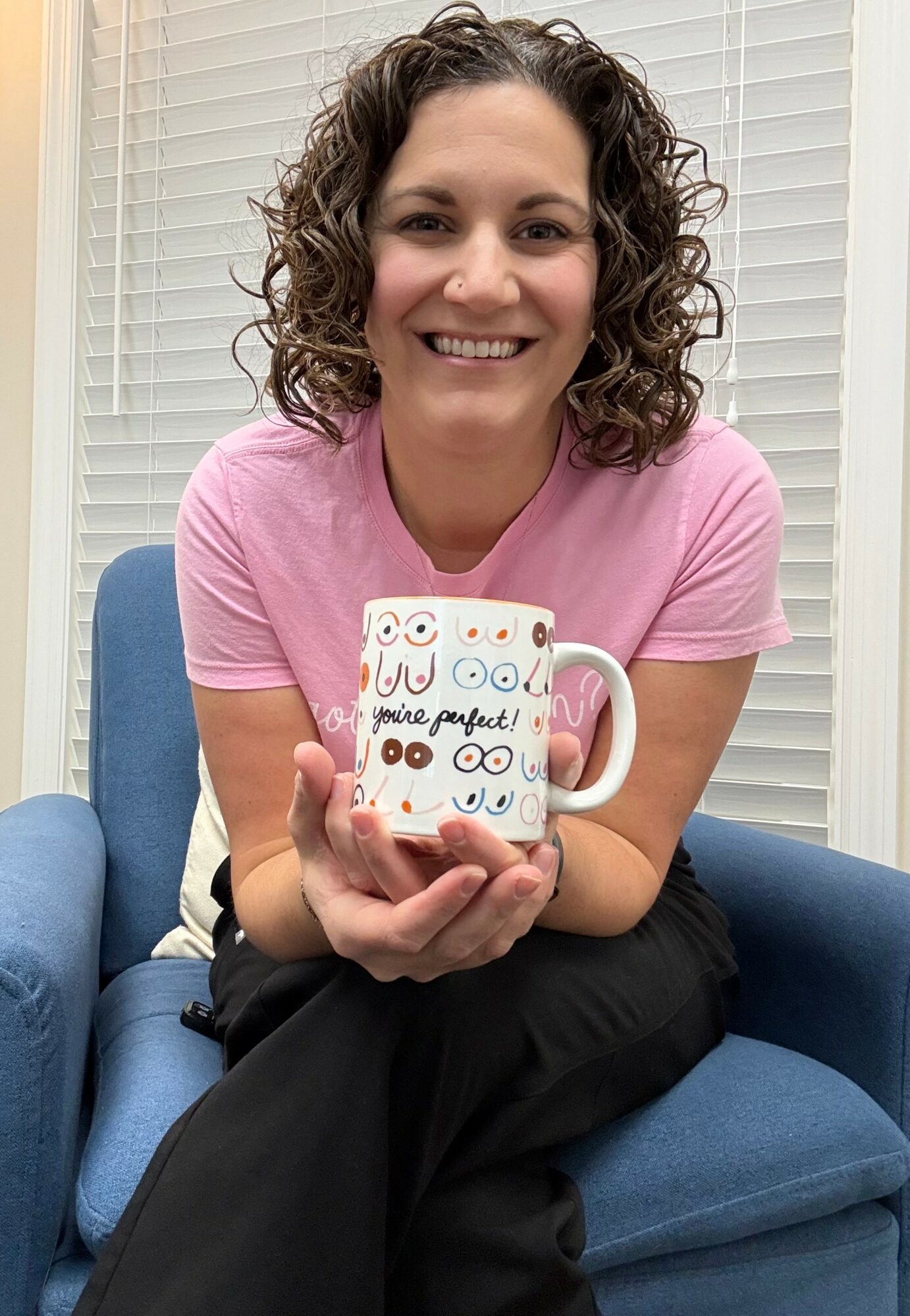 Woman with curly hair smiling, holding a decorated mug, sitting on a blue chair in front of window blinds.
