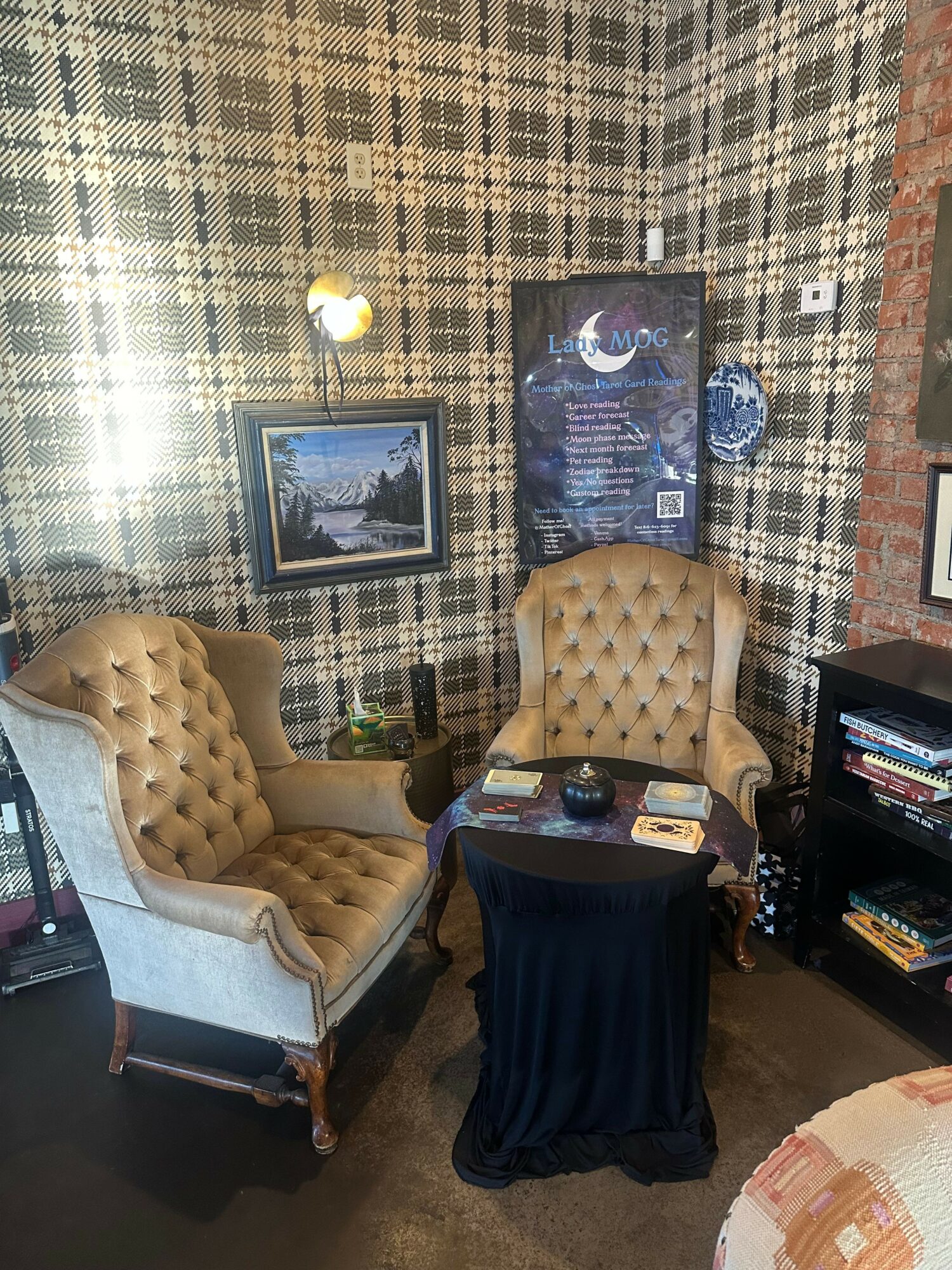 Room with two tufted chairs, a table with a black cloth, bookshelf, framed picture, and patterned wallpaper.