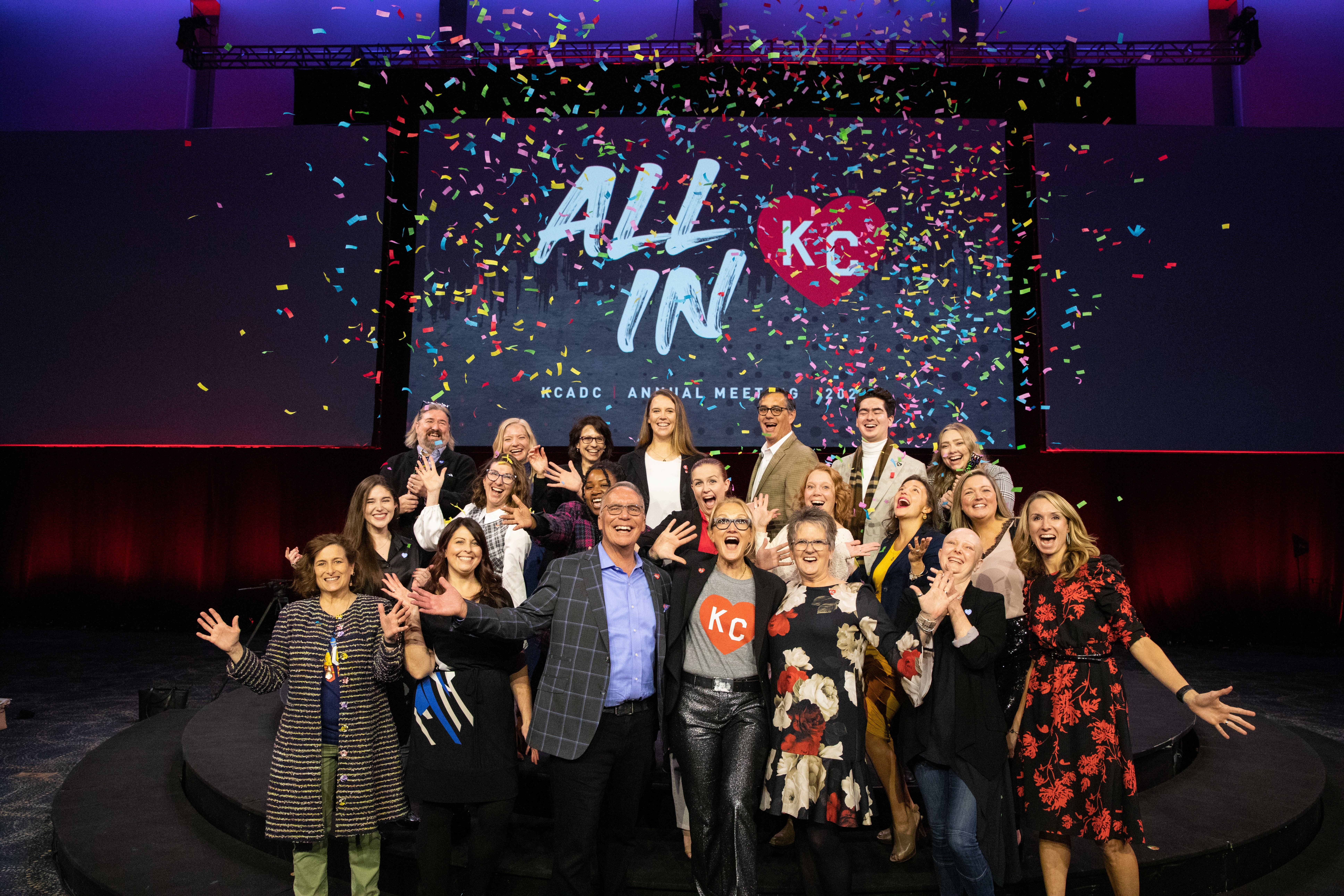 Group of people on stage celebrating with confetti, some raising hands, in front of a screen with text and logo.