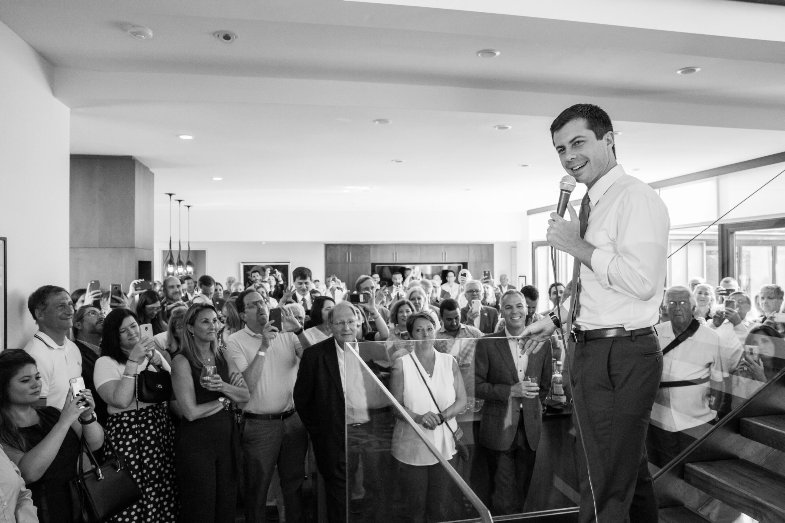 A man stands on a stage speaking to a large crowd in an indoor event space, with people listening attentively.