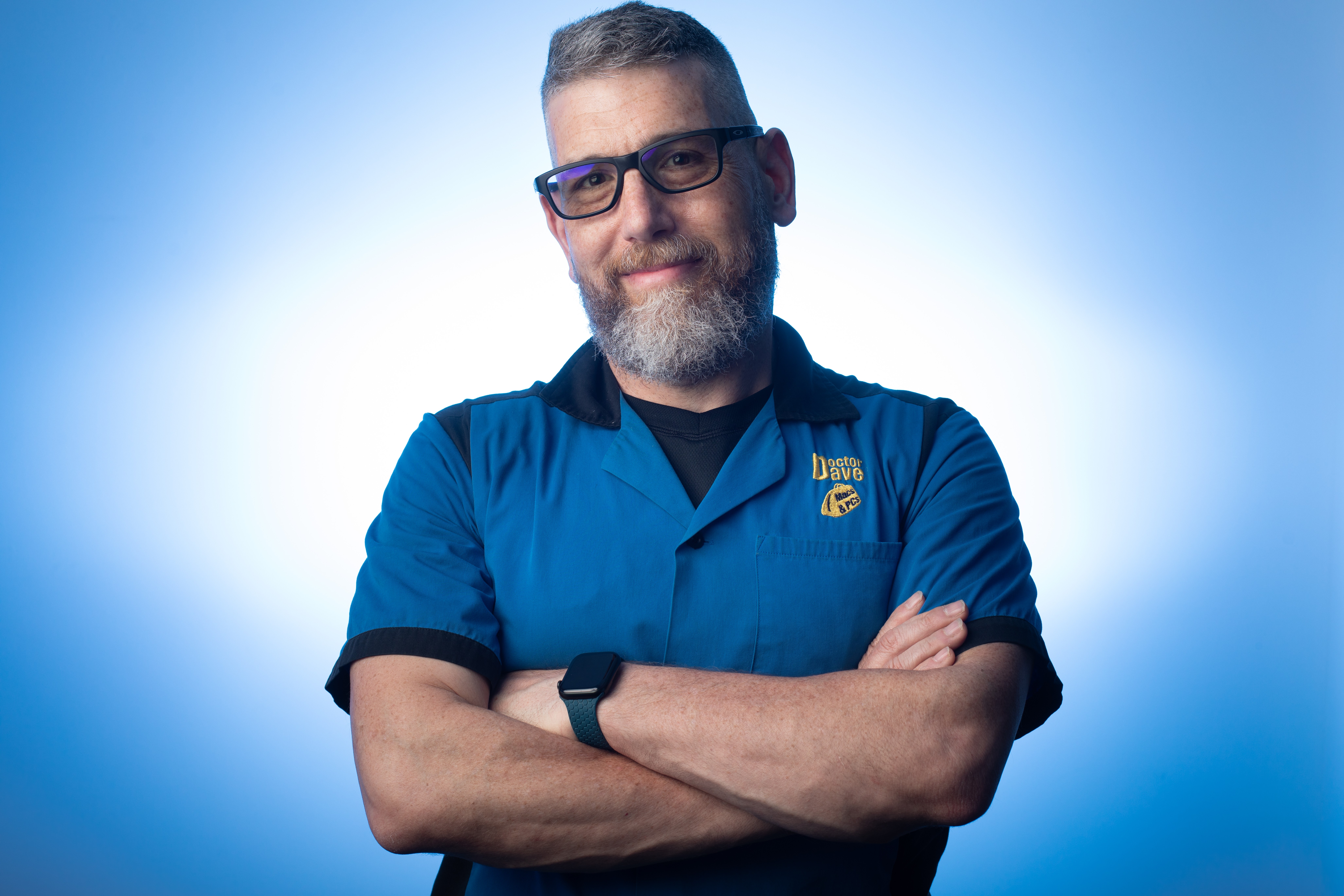 Man with glasses and beard, arms crossed, wearing a blue shirt, smiling, against a blue background.