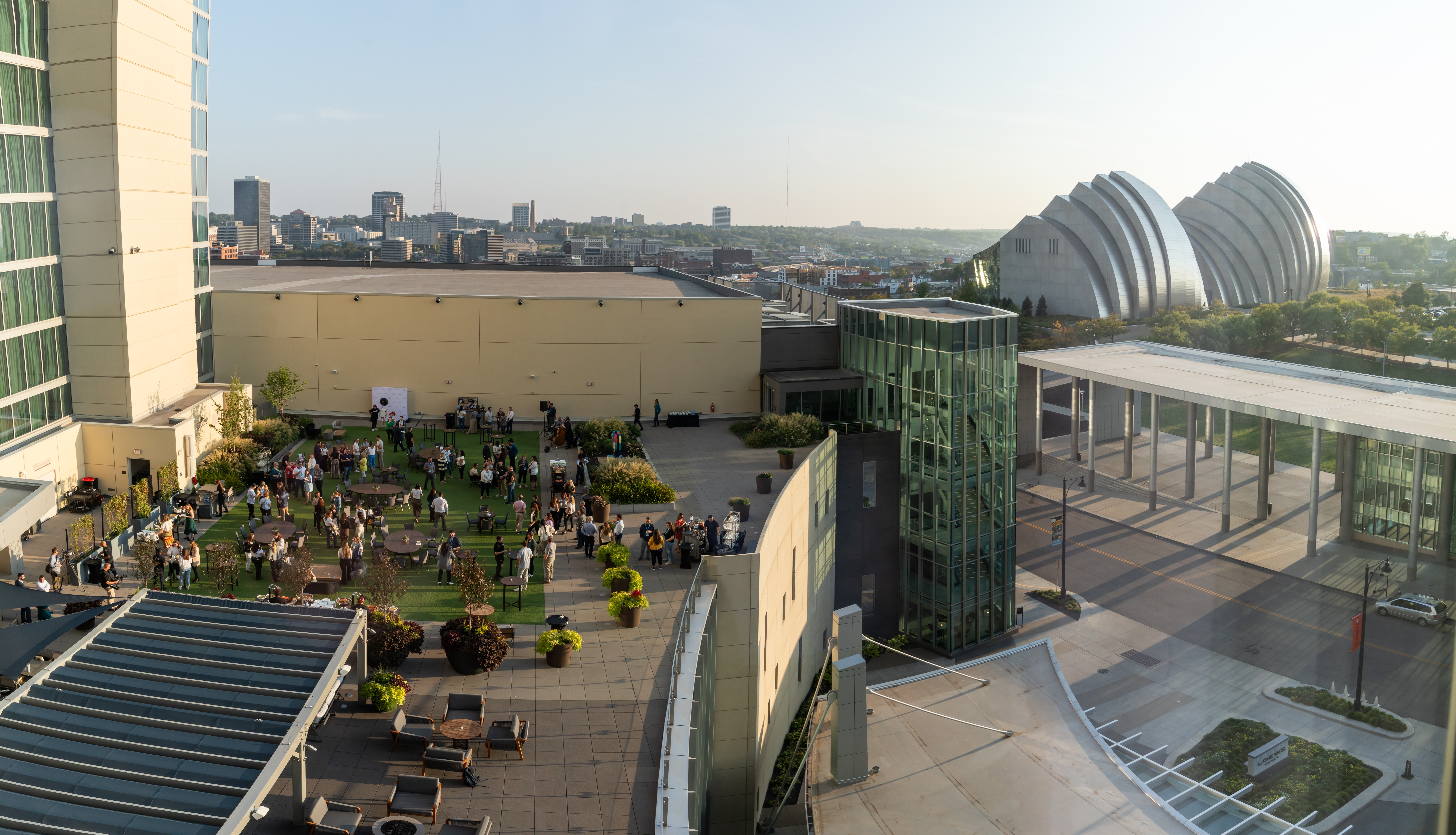 Rooftop terrace with seating and people, city skyline in background, modern buildings, and a glass structure, daytime.