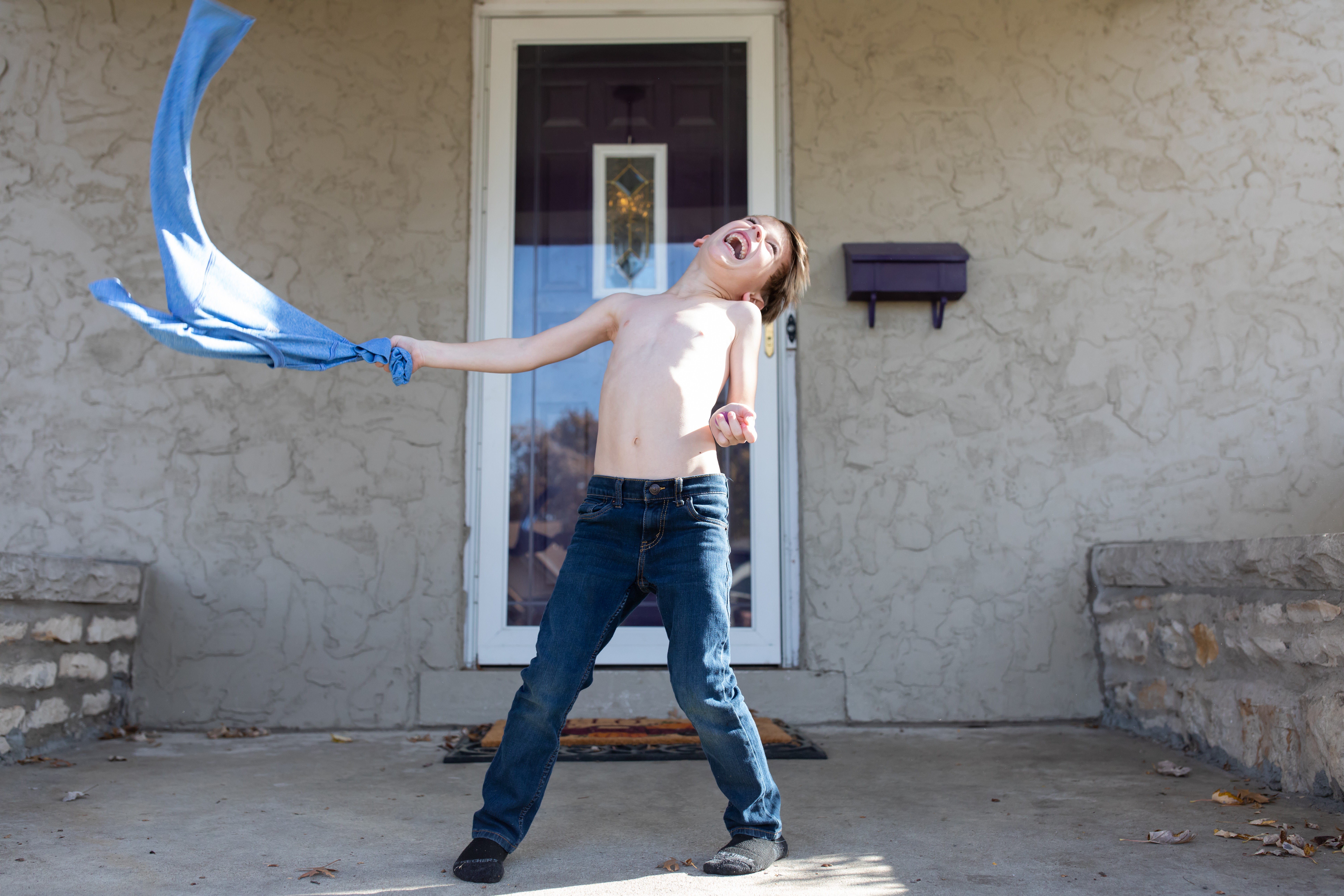 Person standing outside in front of a door, holding a blue cloth, laughing, with arms outstretched and head tilted back.