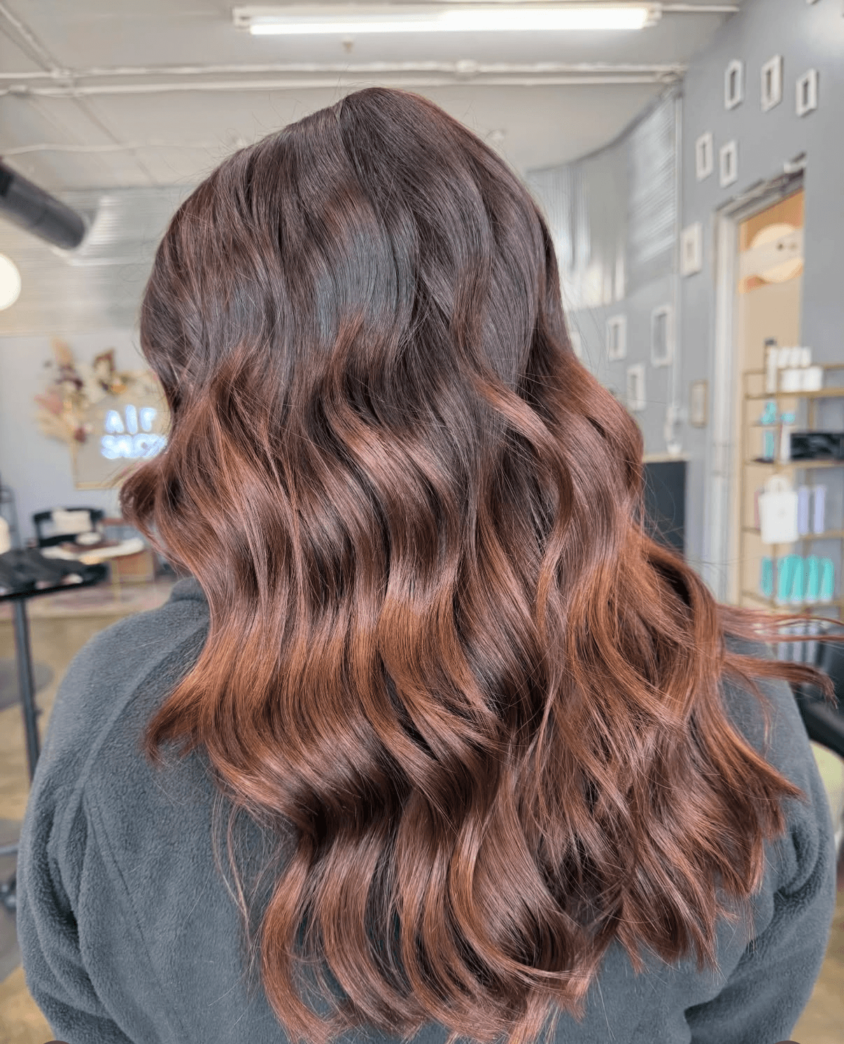 Back of a person with long, wavy, brown hair in a salon, with shelves and mirrors in the background.