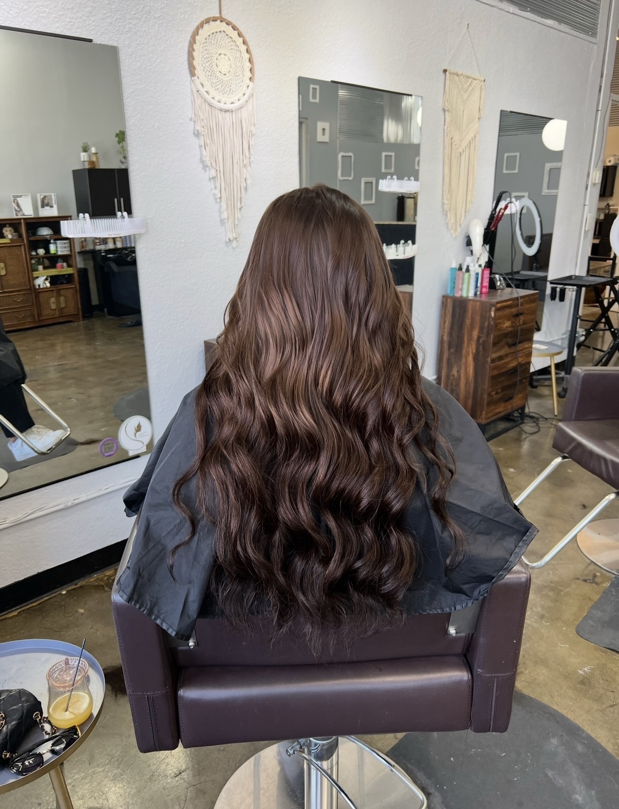 Back view of a person with long, wavy brown hair sitting in a salon chair, facing mirrors and salon equipment.