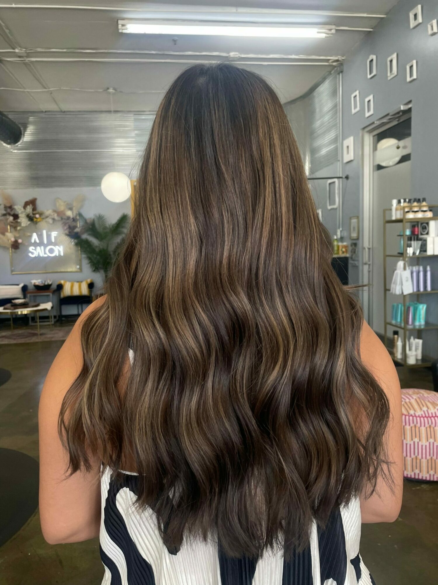 Back view of a person with long, wavy brown hair in a store or salon, shelves and equipment visible in the background.