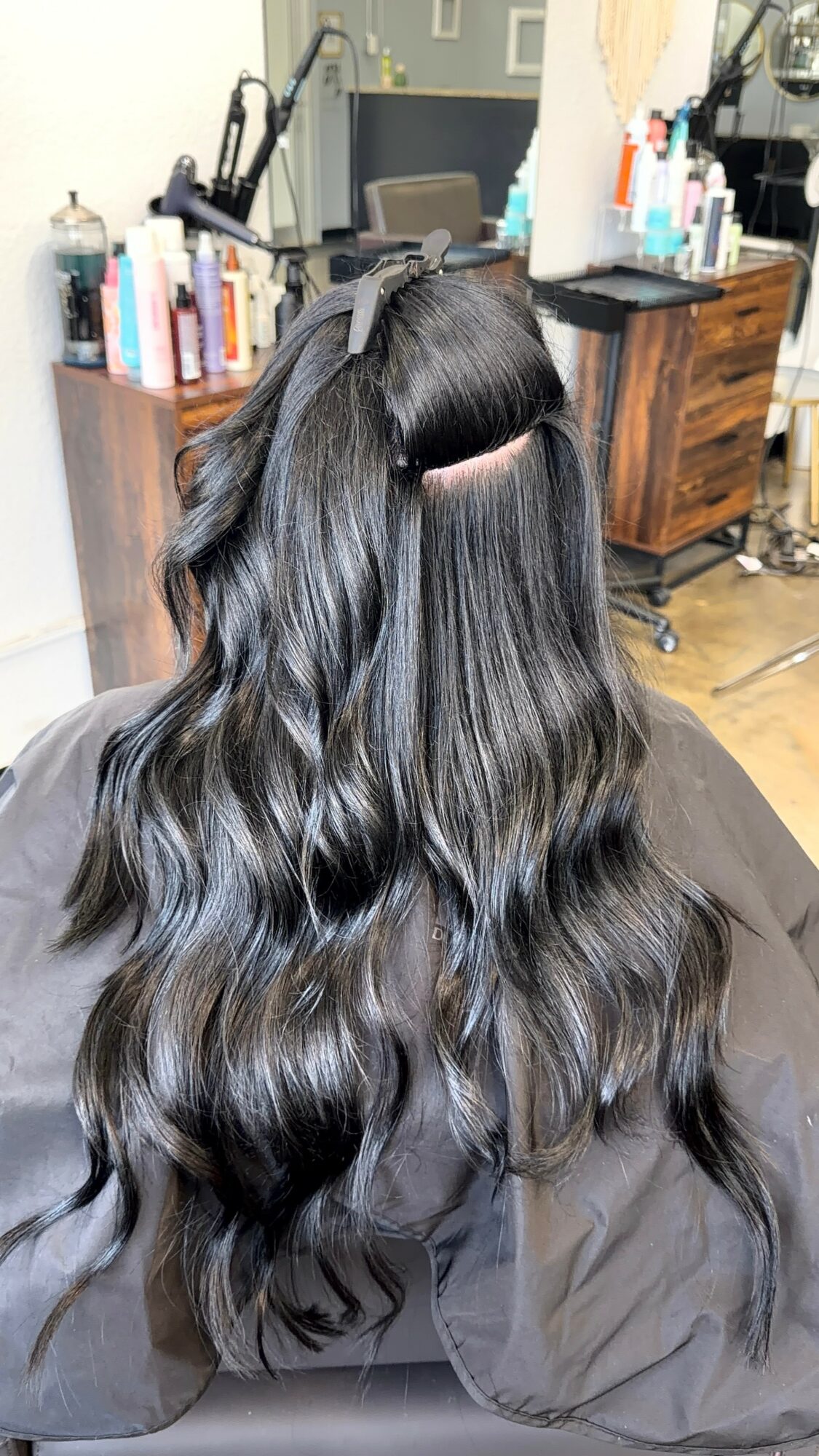 Person with long, wavy black hair sitting in a salon chair, hair sectioned with a clip, salon equipment in background.