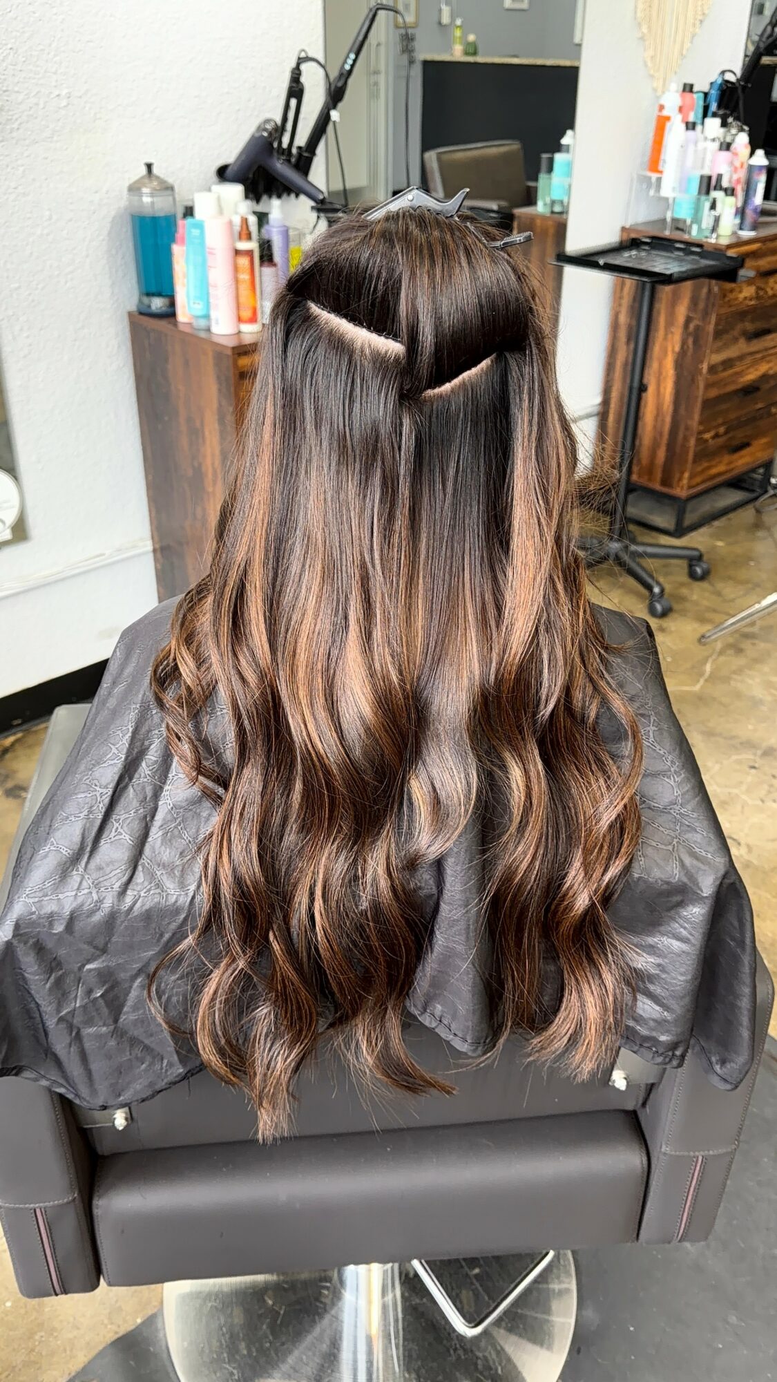 Back view of a woman with long, wavy brown hair in a salon chair, hair sectioned for styling or coloring.