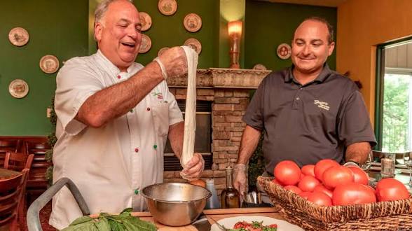 Two men in a kitchen with green and yellow walls, one holding dough, the other smiling near a basket of tomatoes.