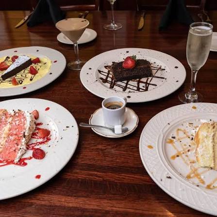 Assorted desserts and drinks on a wooden table, including a chocolate cake, a slice of cake, a mousse, and beverages.