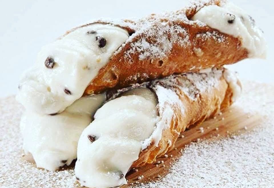 Two cannoli pastries filled with cream and chocolate chips, dusted with powdered sugar, on a wooden surface.