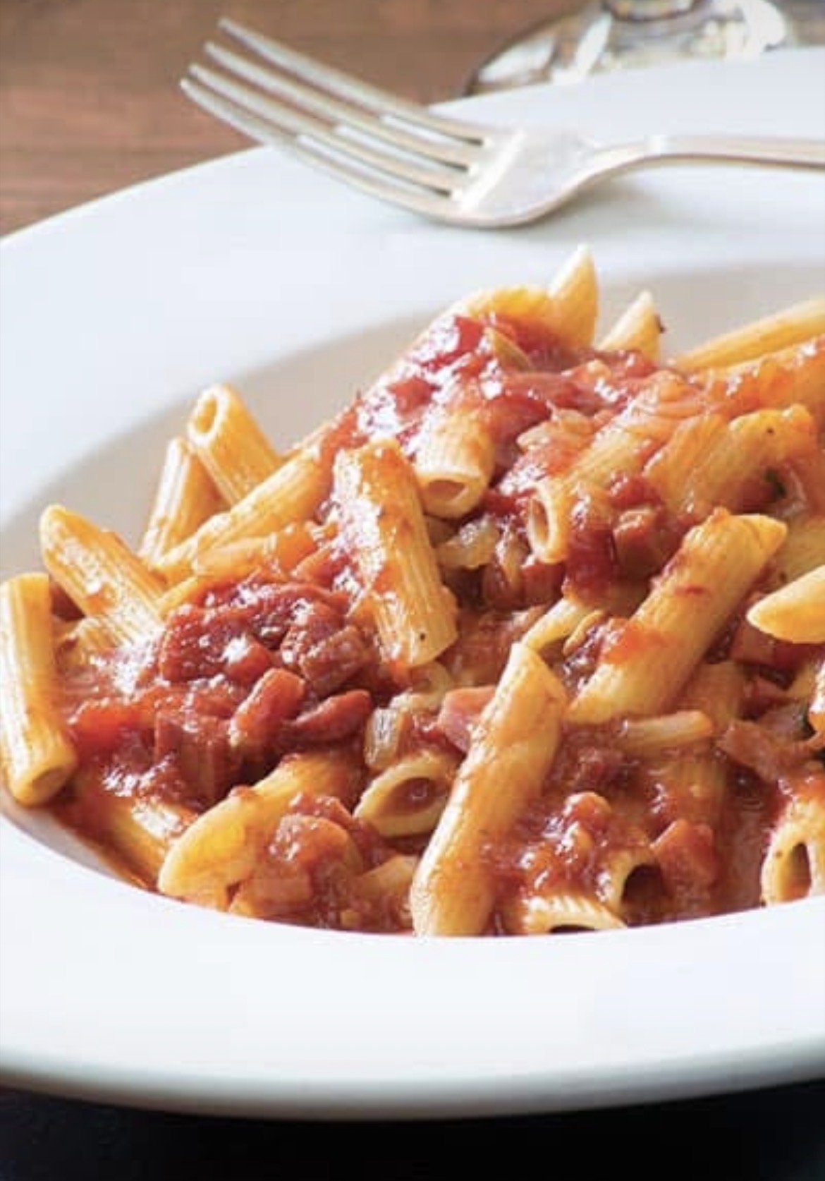 Plate of pasta with tomato sauce on a white dish, fork and knife in the background.