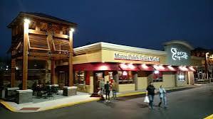 Night scene of a shopping center with a wooden tower structure and a store with a red awning, people walking outside.