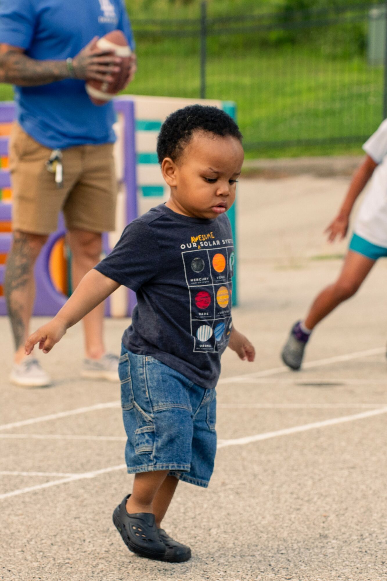 Young boy walking on a sports court holding a ball, with other children and a person in the background.