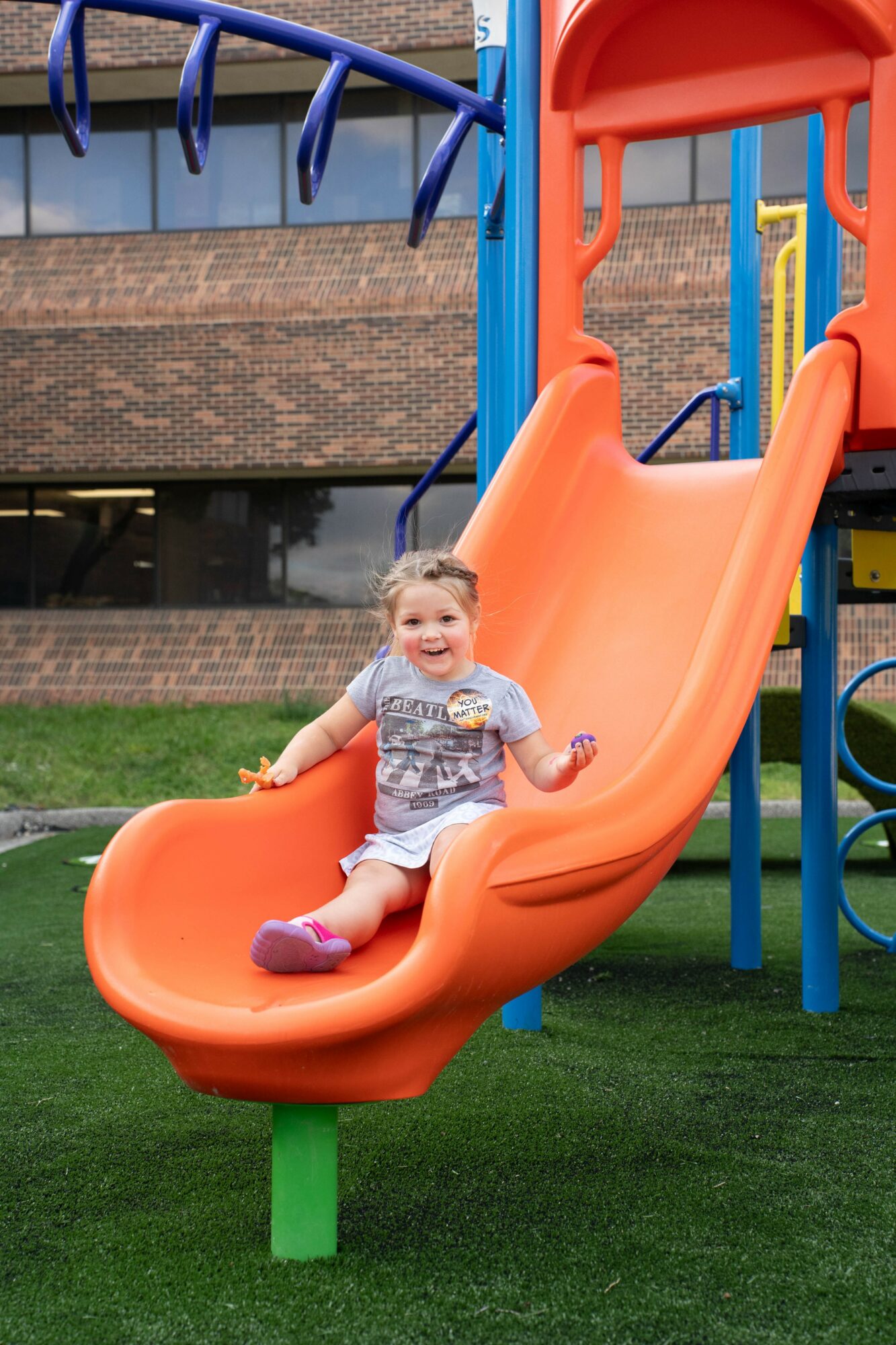 Child smiling on orange slide at playground, holding sides, with building in background.