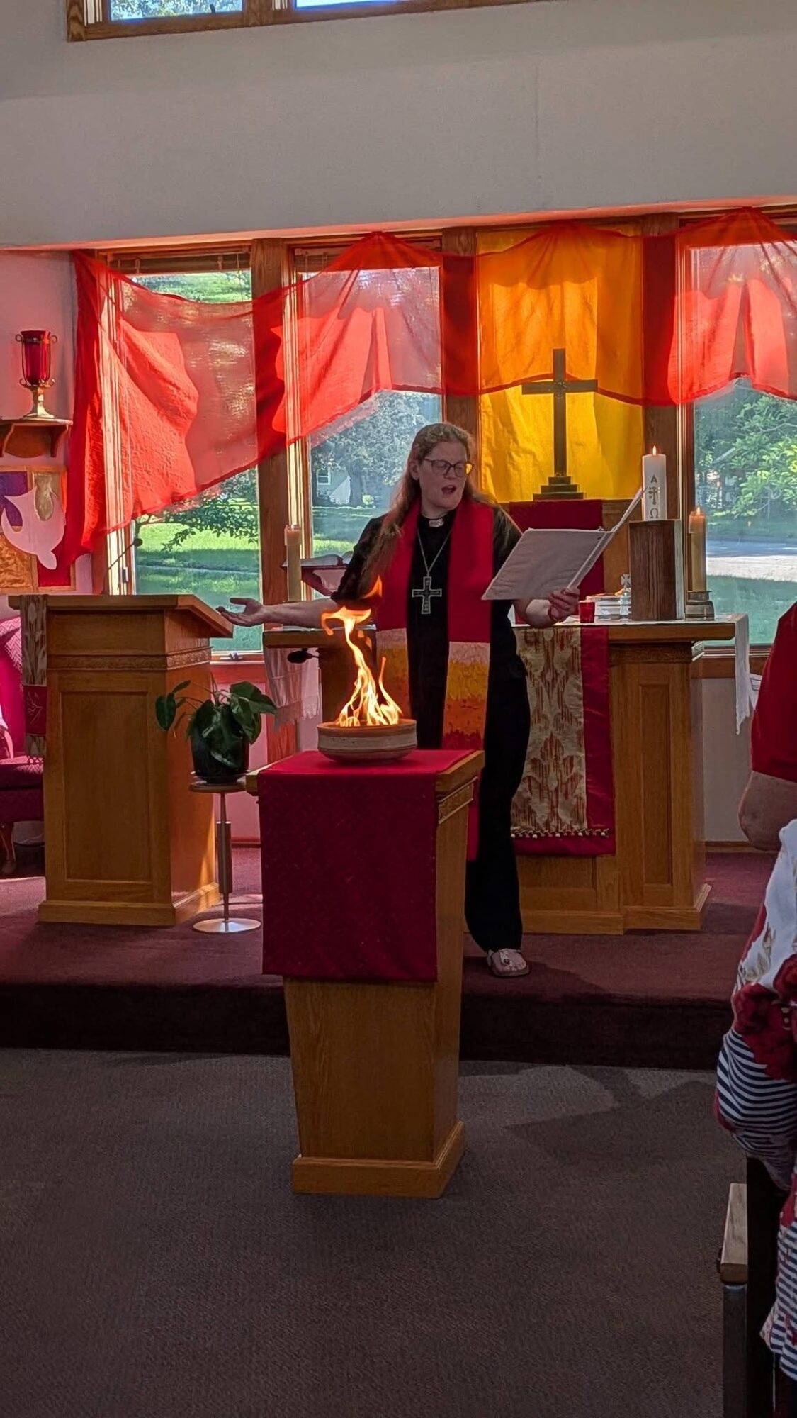 Woman standing at a lectern inside a decorated room with red and orange curtains, holding a paper, with a cross and window behind.